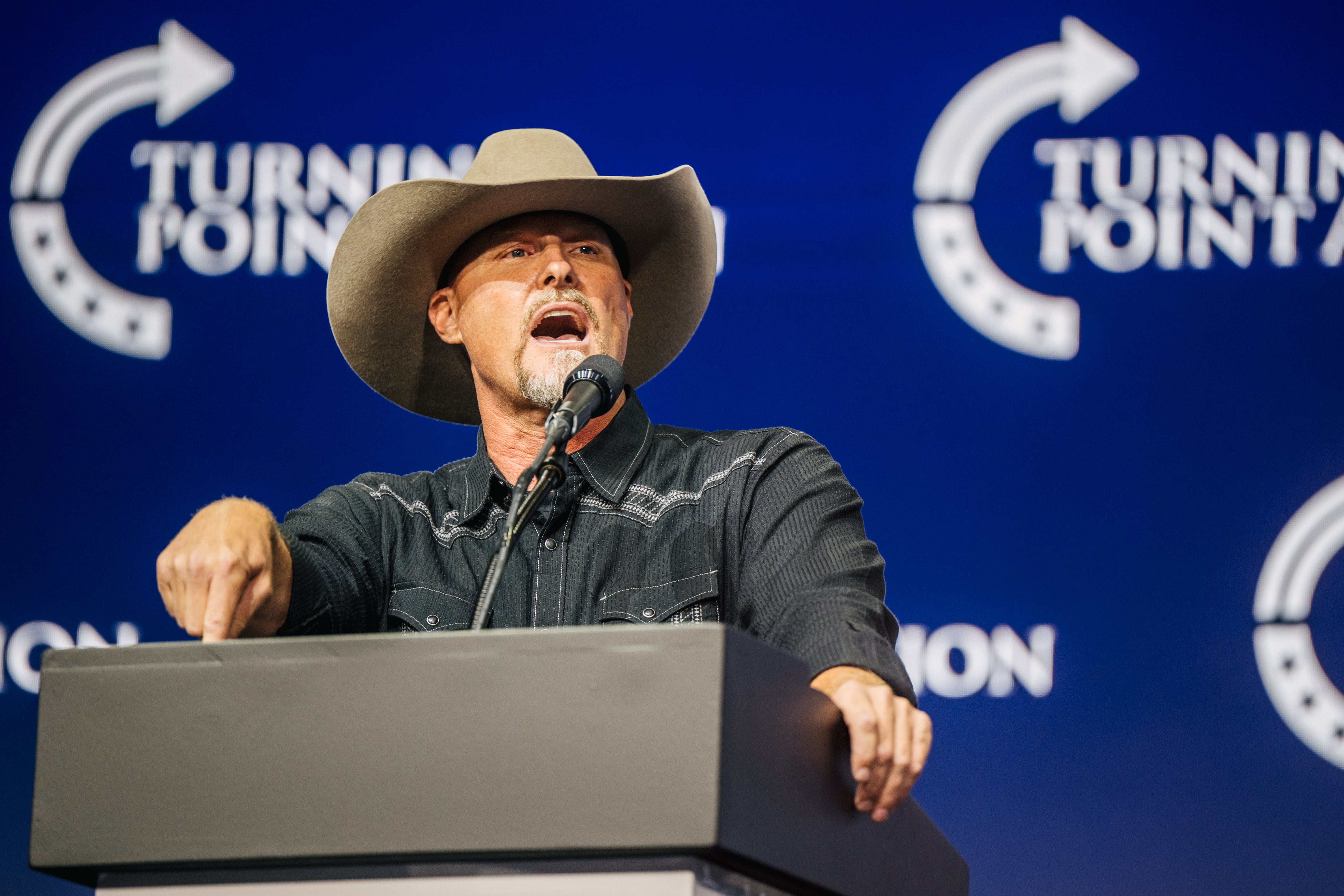 A bearded man in a dark western-style shirt and cowboy hat gestures at a podium against a dark blue backdrop with “Turning Point” written on it. 