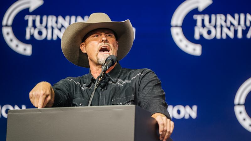 A bearded man in a dark western-style shirt and cowboy hat gestures at a podium against a dark blue backdrop with “Turning Point” written on it.&nbsp;