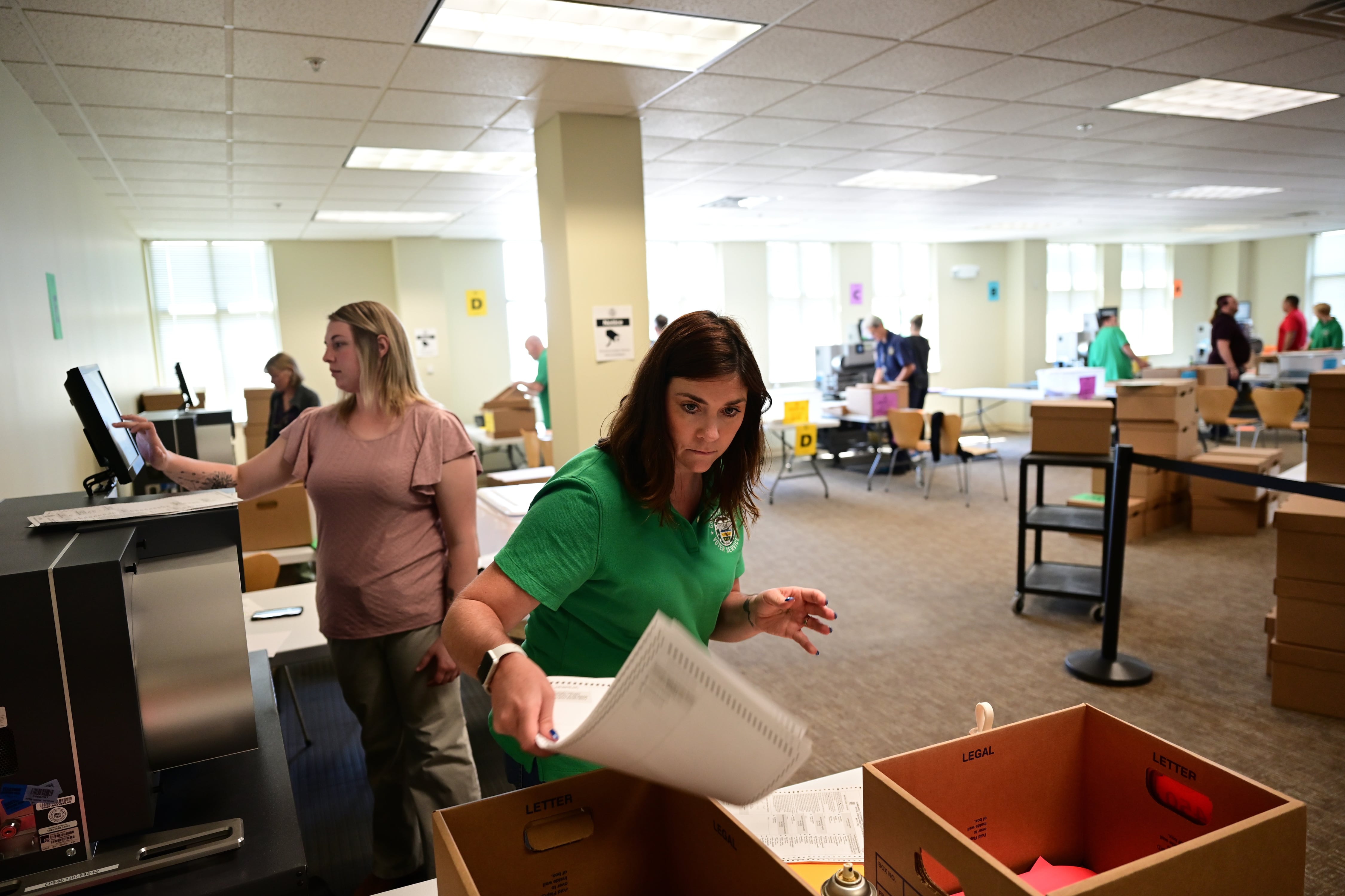 A woman places ballots into a box in a room full of workers 