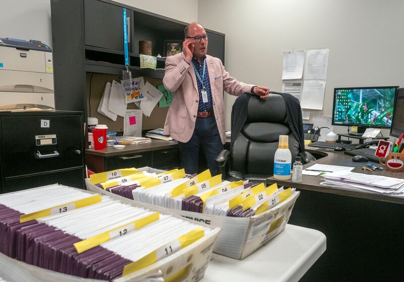 A man wearing a red and white striped suit stands behind a desk holding a phone to his ear. There two boxes of ballots in envelops on the table.
