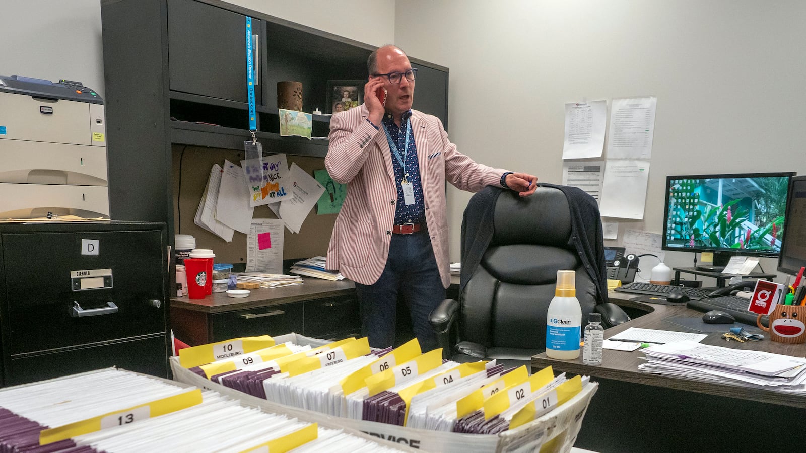 A man wearing a red and white striped suit stands behind a desk holding a phone to his ear. There two boxes of ballots in envelops on the table.