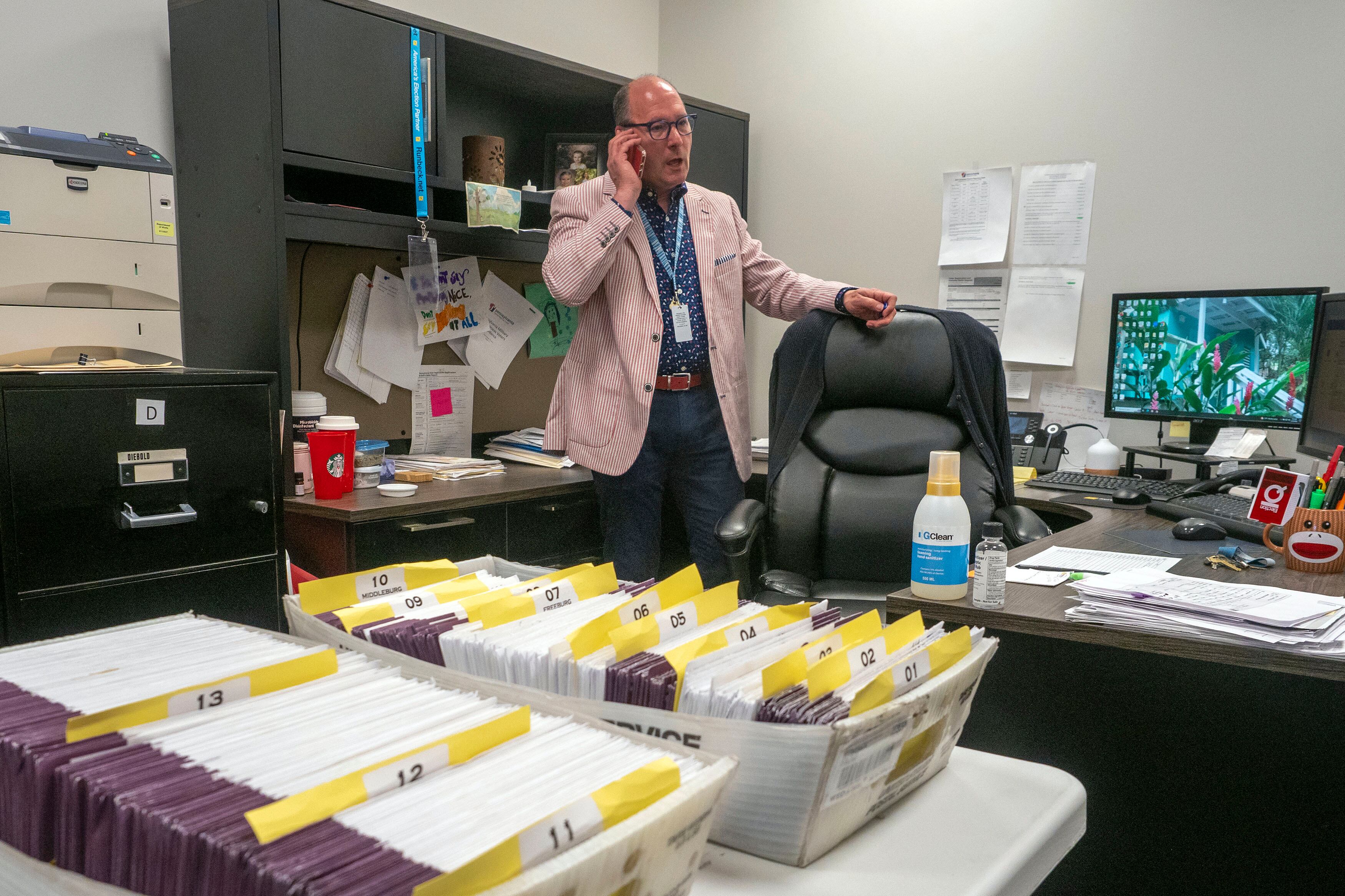A man wearing a red and white striped suit stands behind a desk holding a phone to his ear. There two boxes of ballots in envelops on the table.