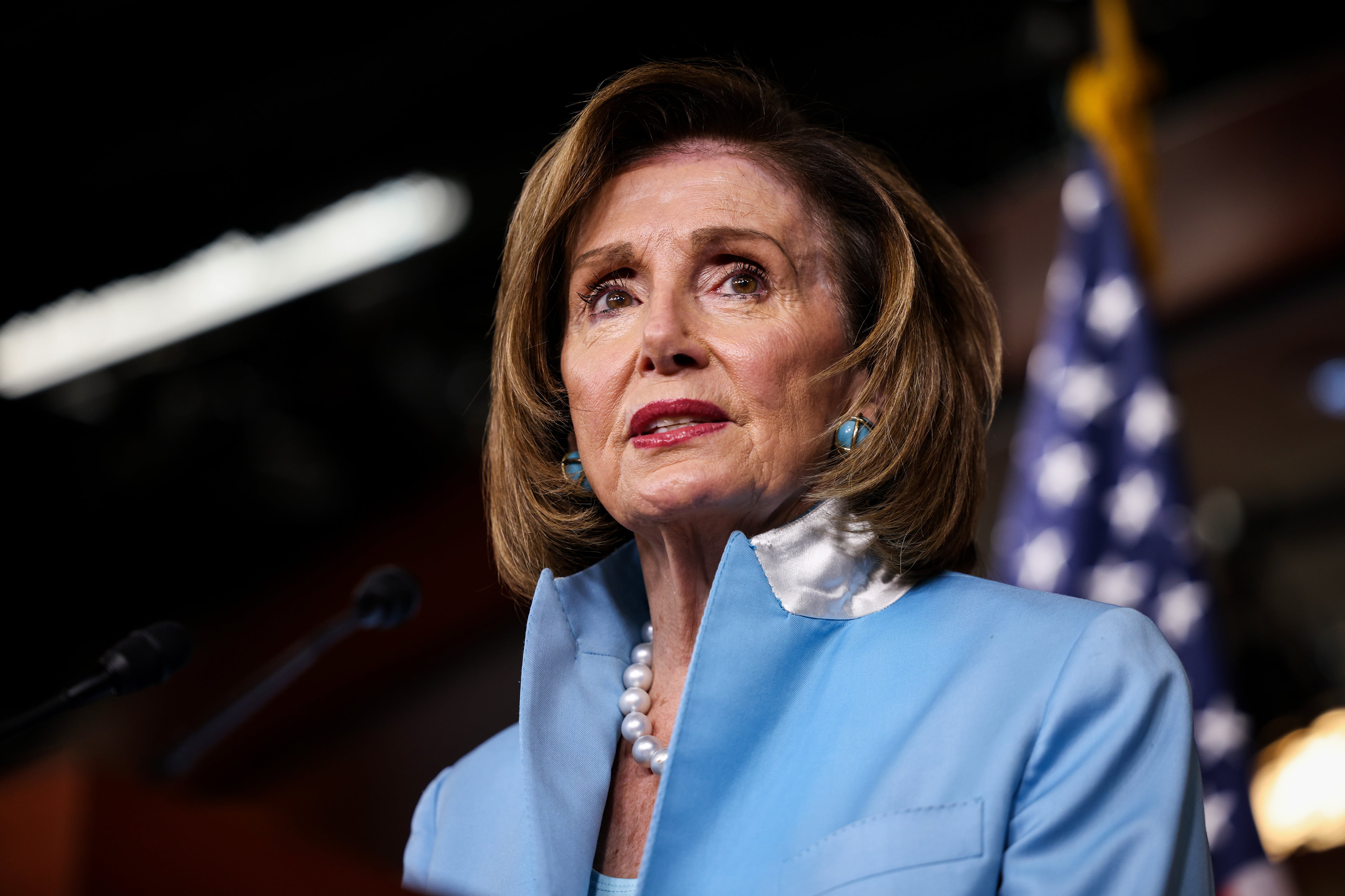 Nancy Pelosi in a light blue jacket looking into the distance with a U.S. flag behind her.