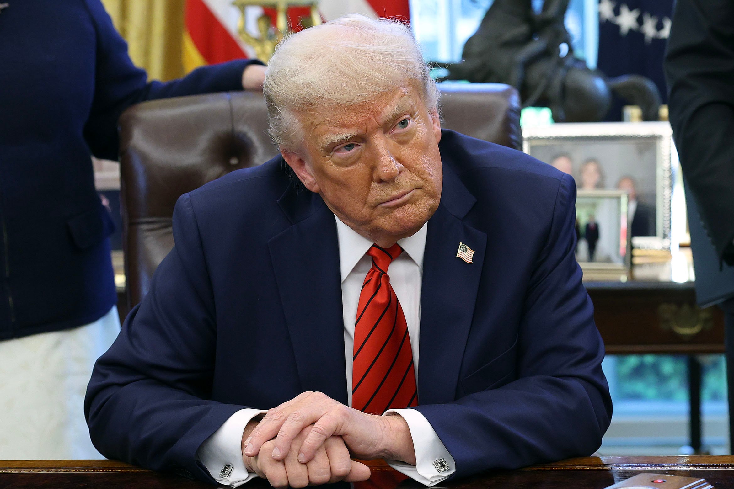 President Trump Signs Executive Orders In The Oval Office while crossing his hands sitting at a wooden desk.