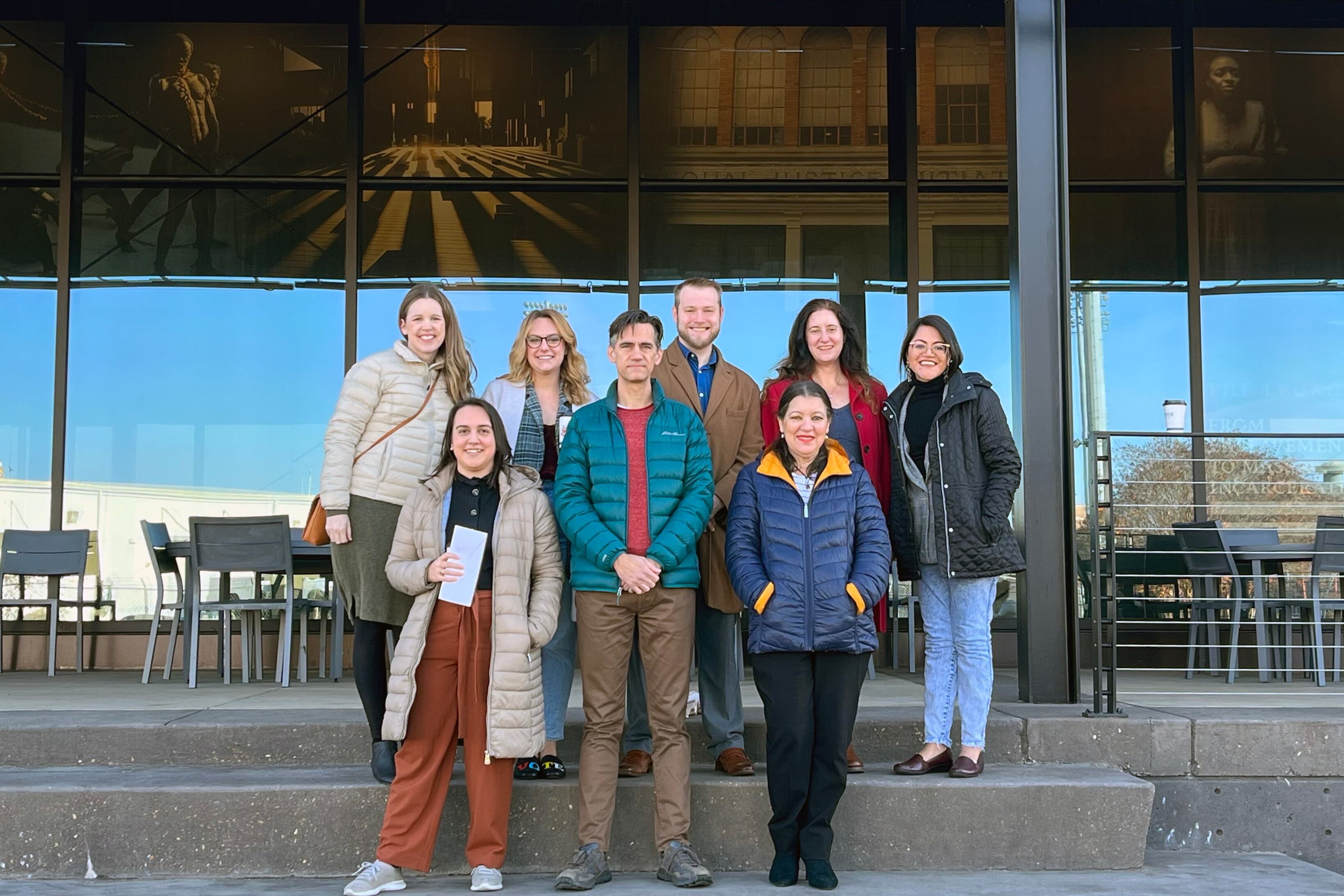 A group of eight people pose for a portrait while standing on stairs in front of a glass building.