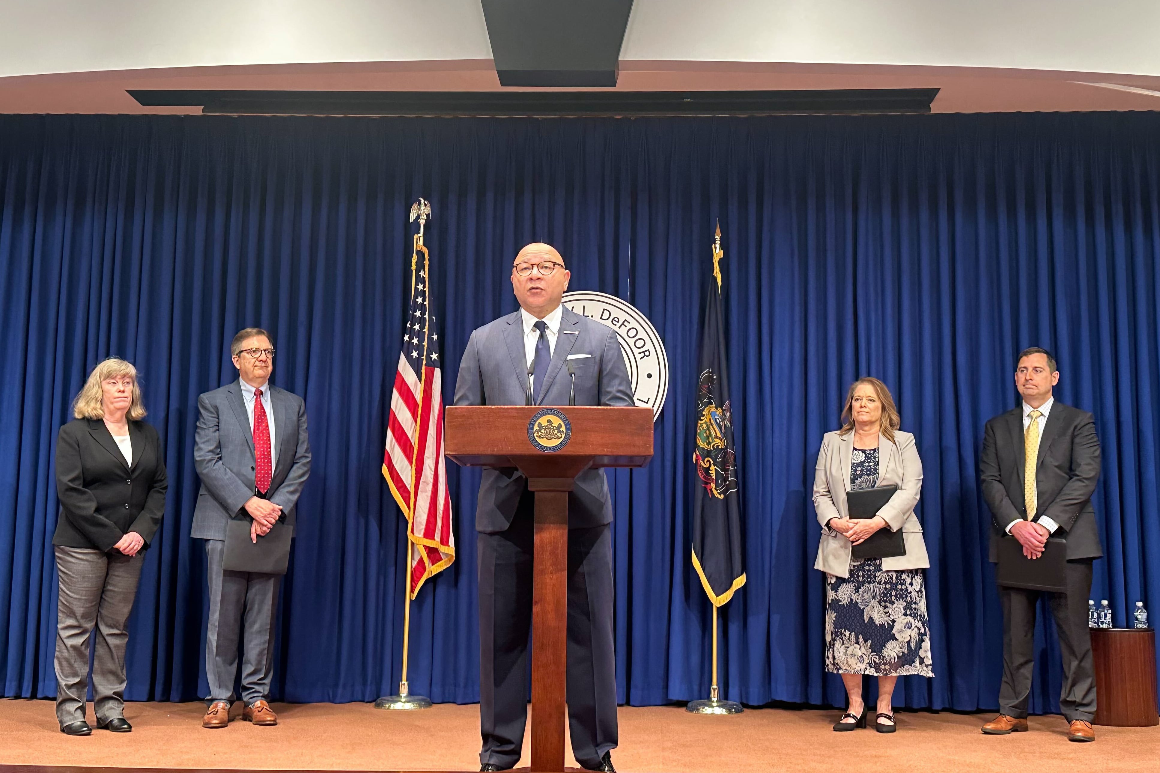 A photograph of a man in a suit speaking from a wooden podium on stage while four men and women in business clothes stand in the background.