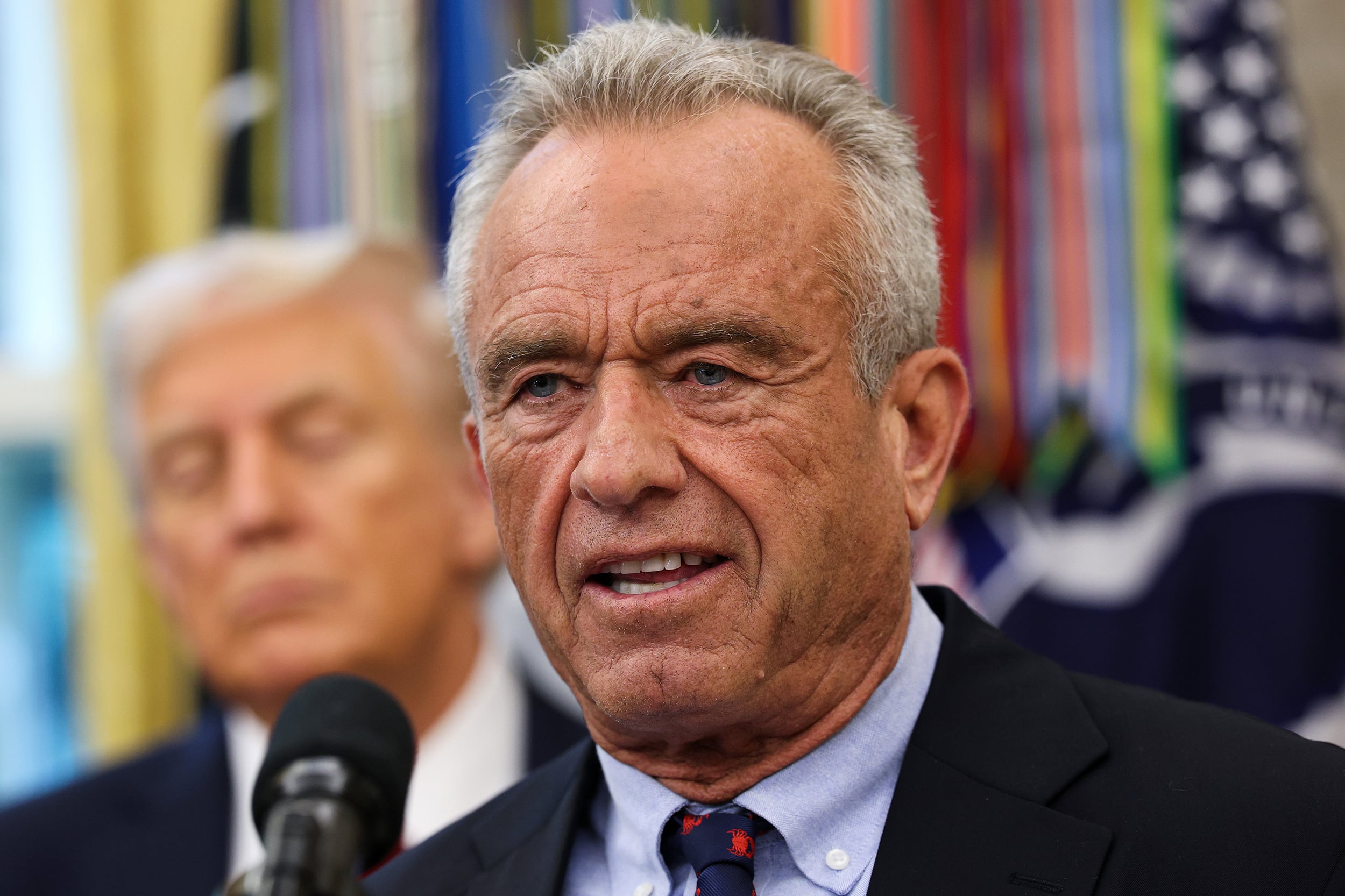 A photograph of an older white man in a suit speaking from a microphone with President Trump in the background.