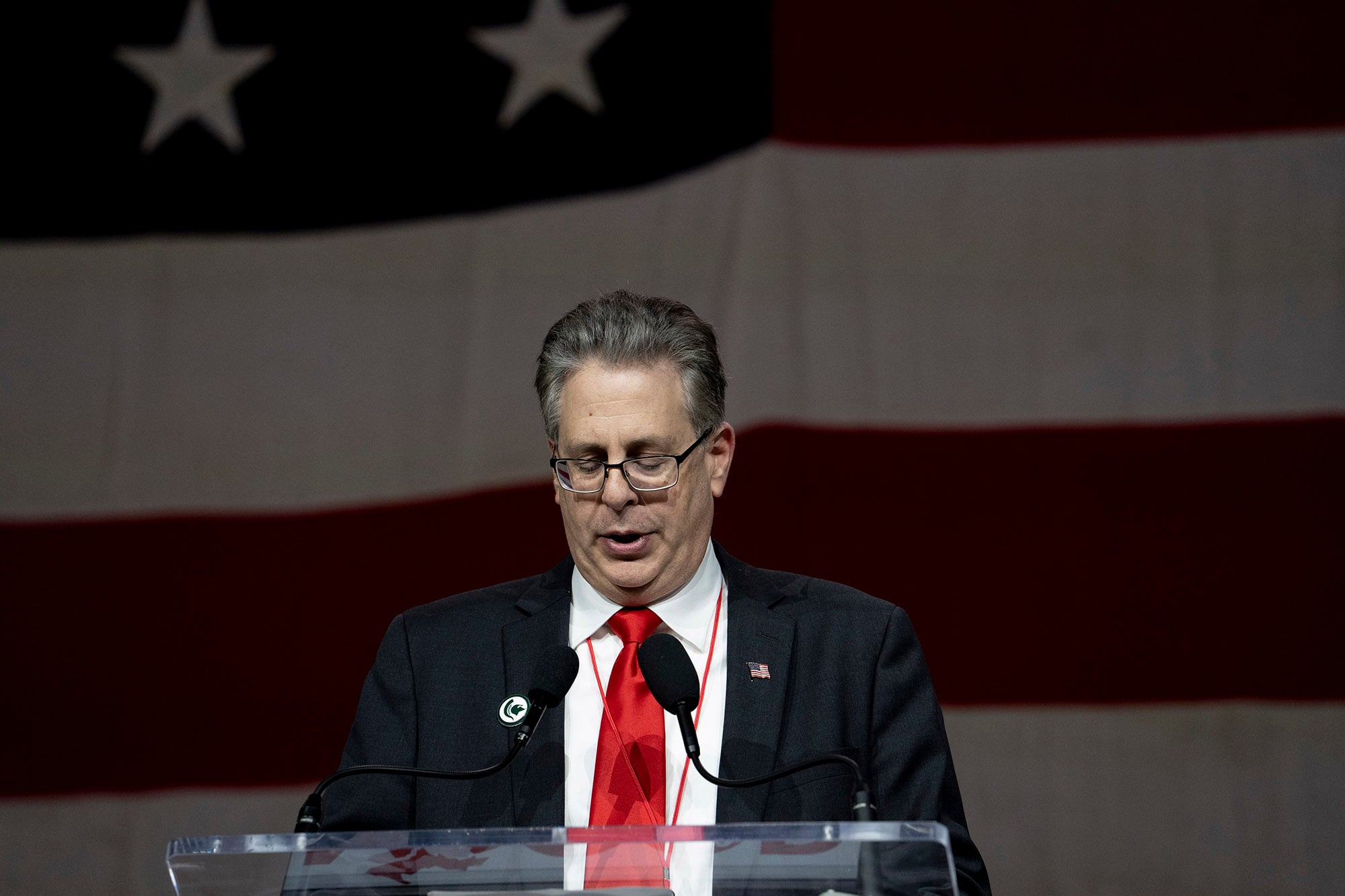 A photograph of a white man in a suit stands behind a podium and in front of a large American flag.
