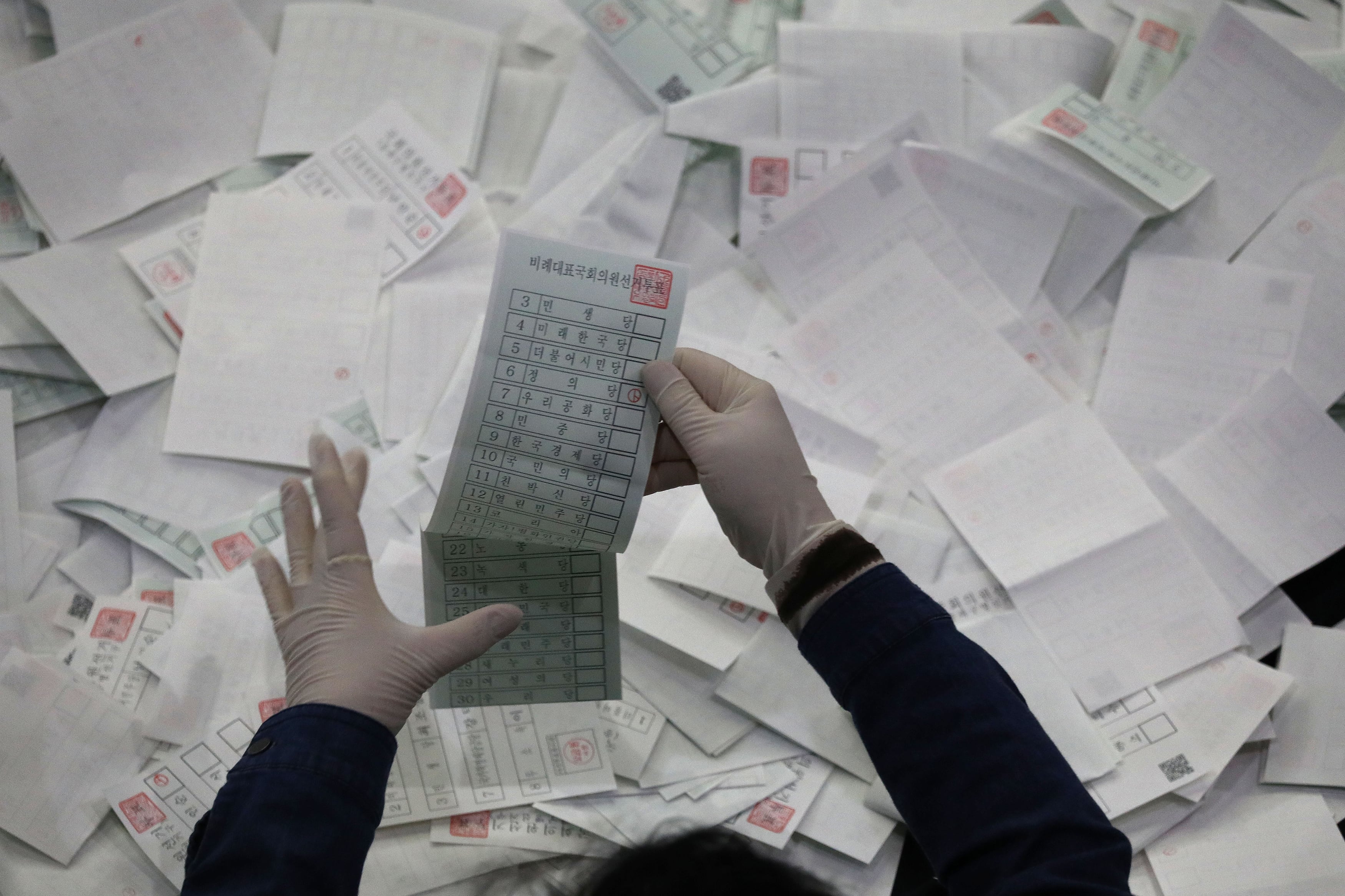 An aerial view of a pair of hands in gloves holding a ballot over the top of a giant pile of ballots.