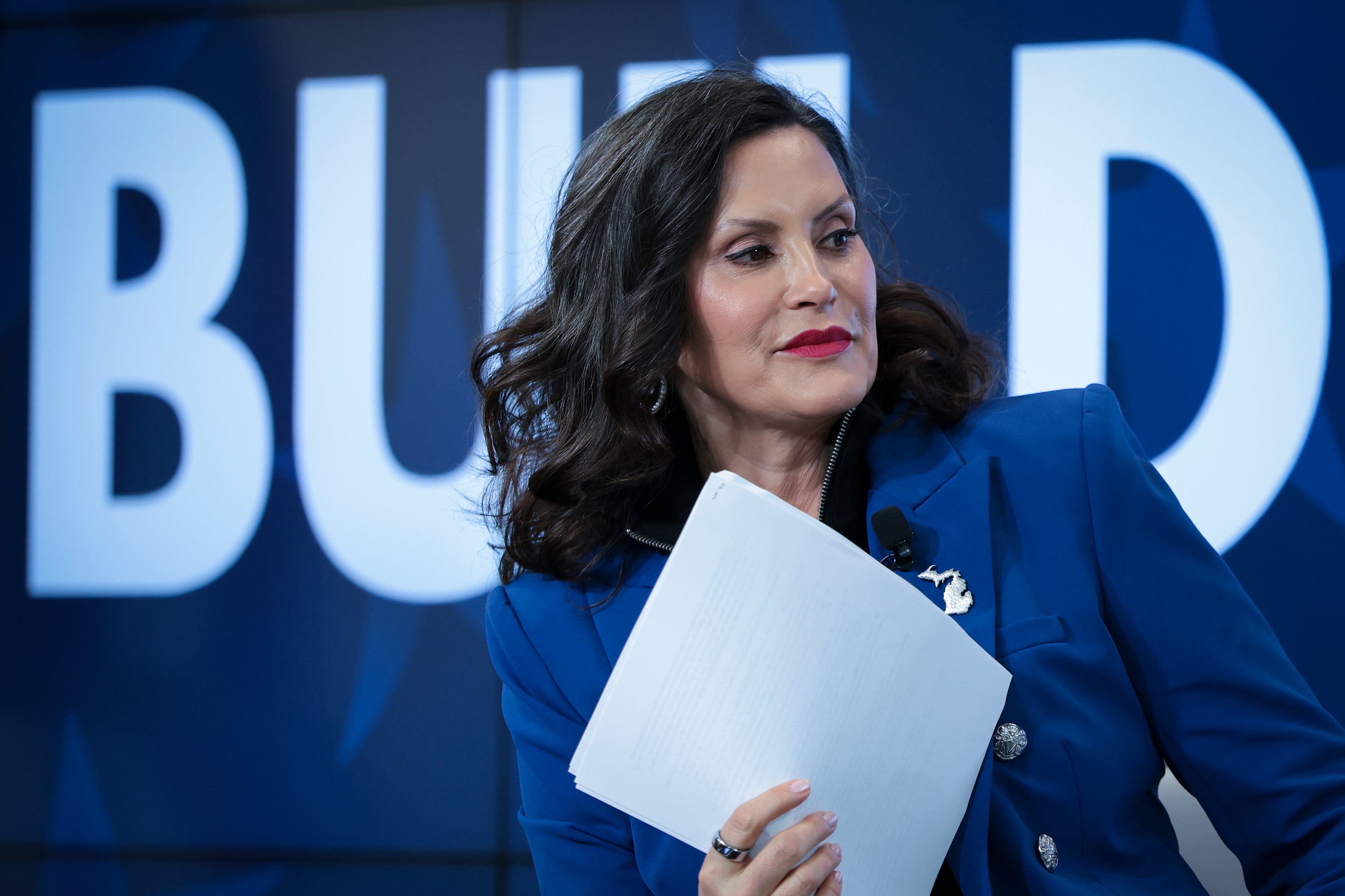 A photograph of a white woman in a blue suit holding a pile of paper in her hand.