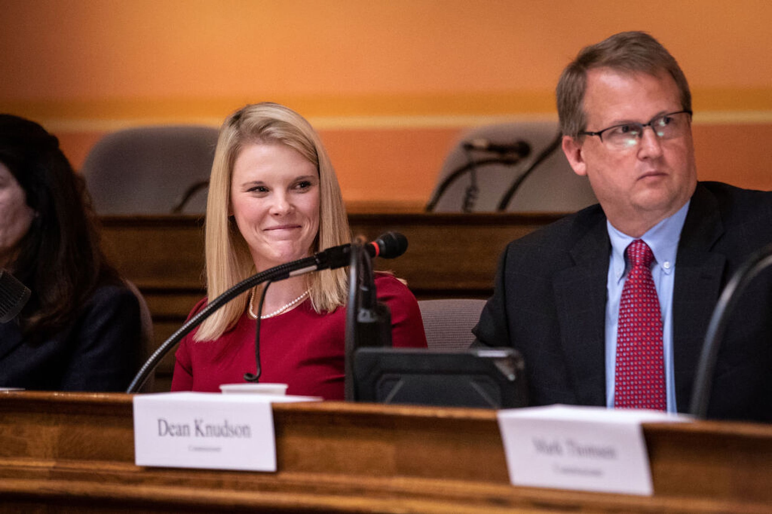 Two people in business clothes sit next to each other behind microphones and behind wooden desk.