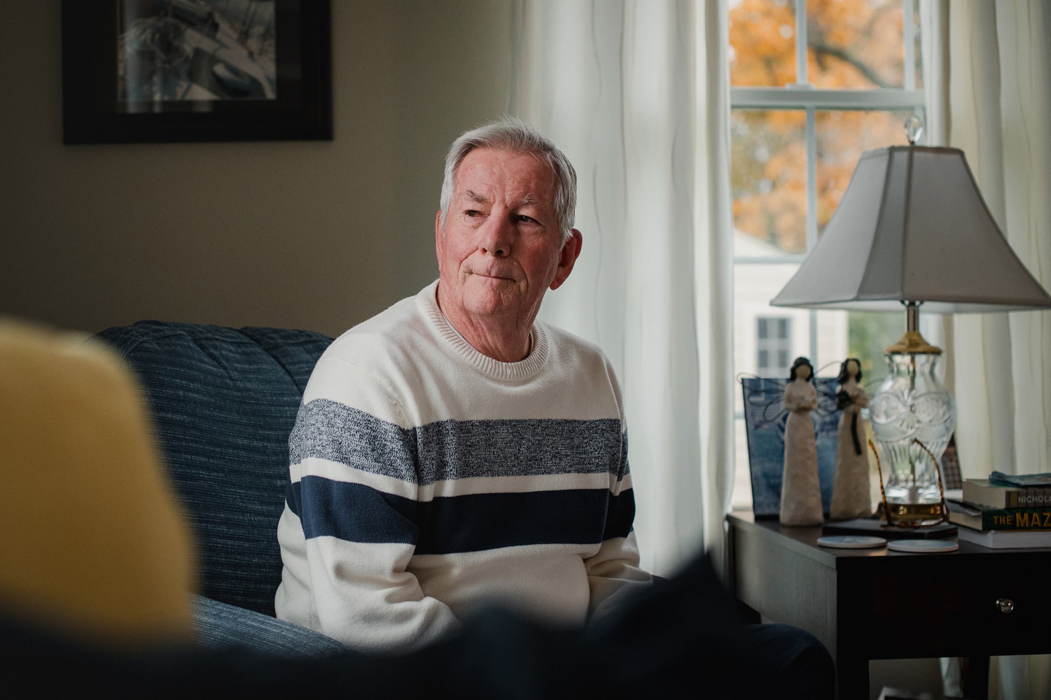 A man with white hair and wearing a white sweater with two blue stripes sits on a chair next to a lamp with a window in the background.
