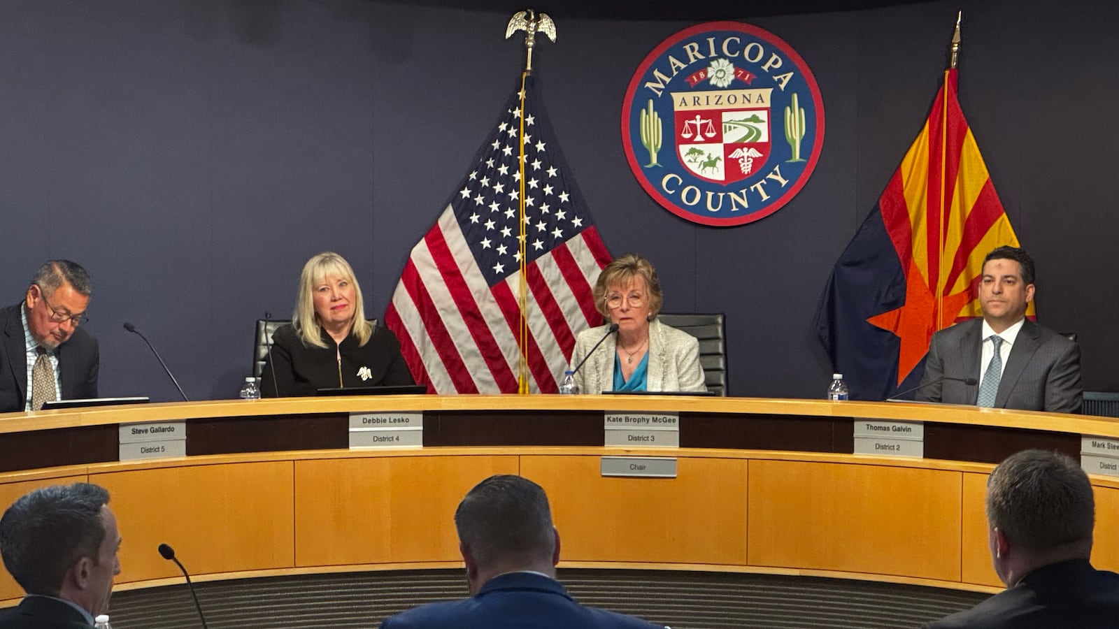 A photograph of a group of people in suits sitting at a wooden table with flags and facing a group of people sitting in chairs all in a conference room.