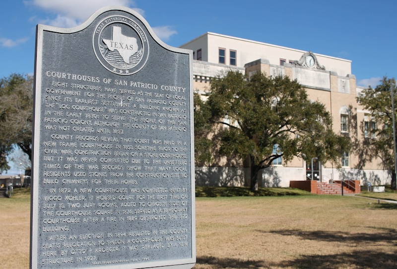 A grey plague with a historical description in the foreground with a large building and trees in the background.