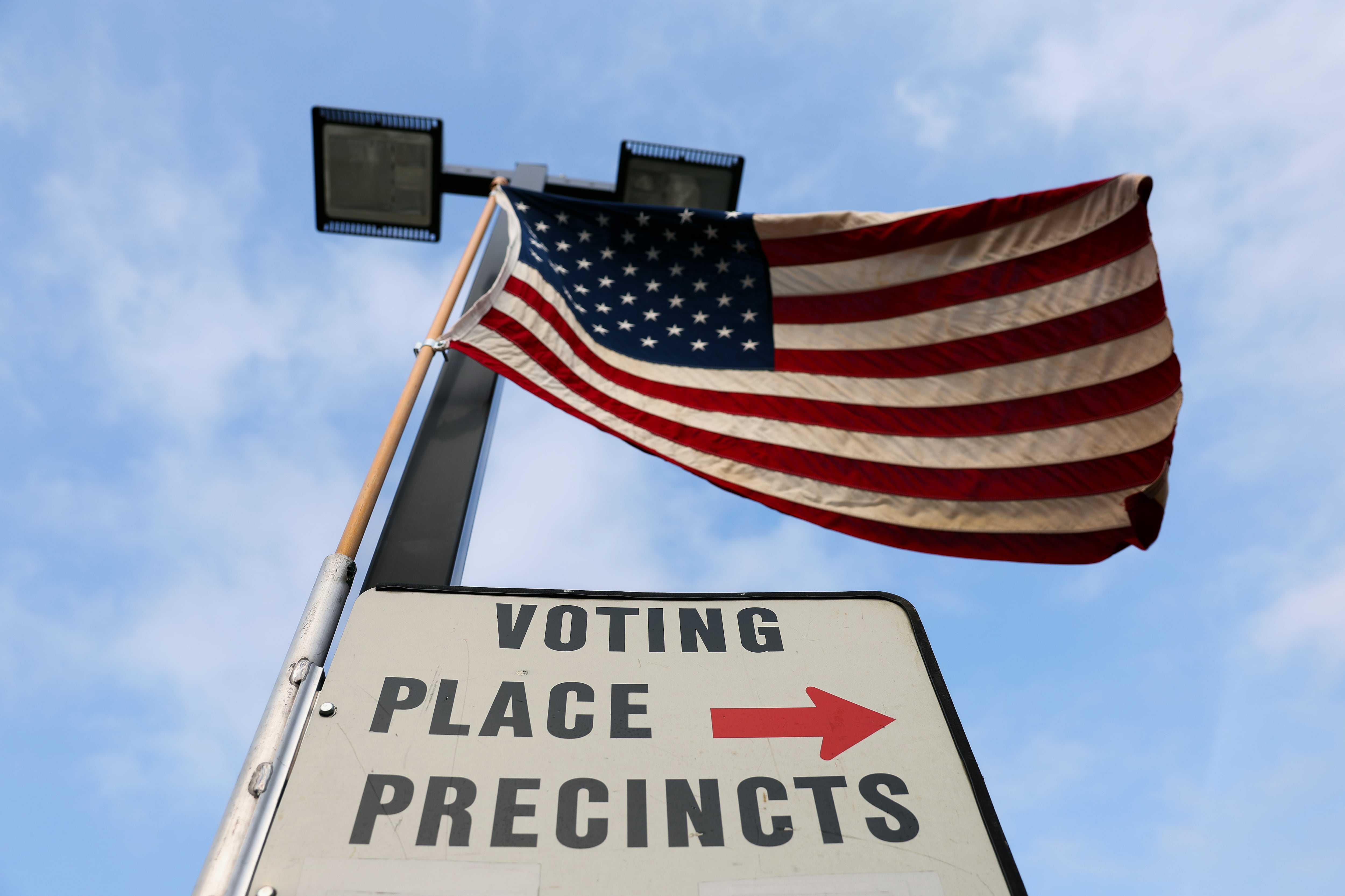 A U.S. flag waves above an outdoor sign reading "voting place precincts" with an arrow pointing right