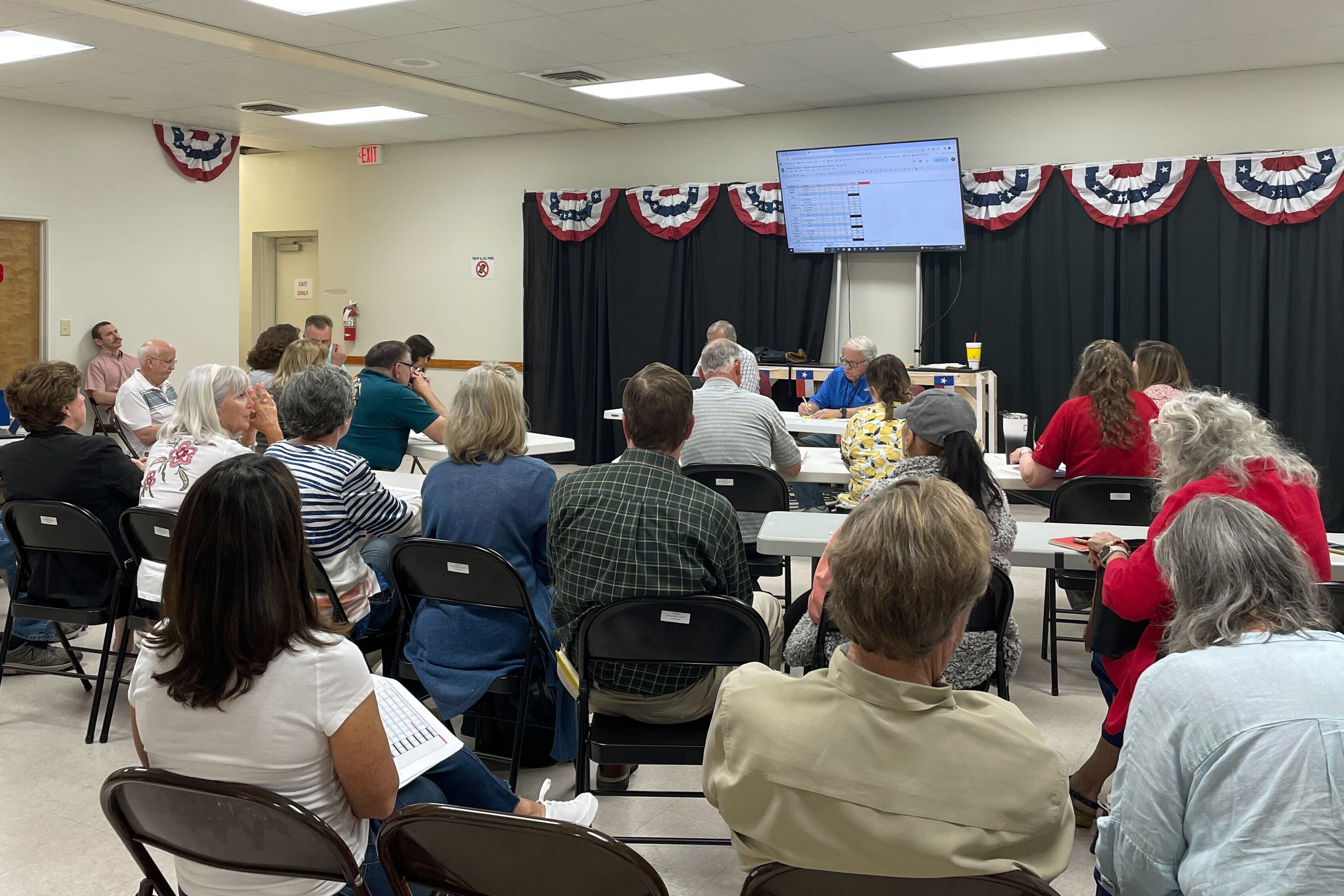 Two people sit at a small, white folding table in front of a large group of people sitting in chairs in rows. There is a TV and a dark backdrop in the background.