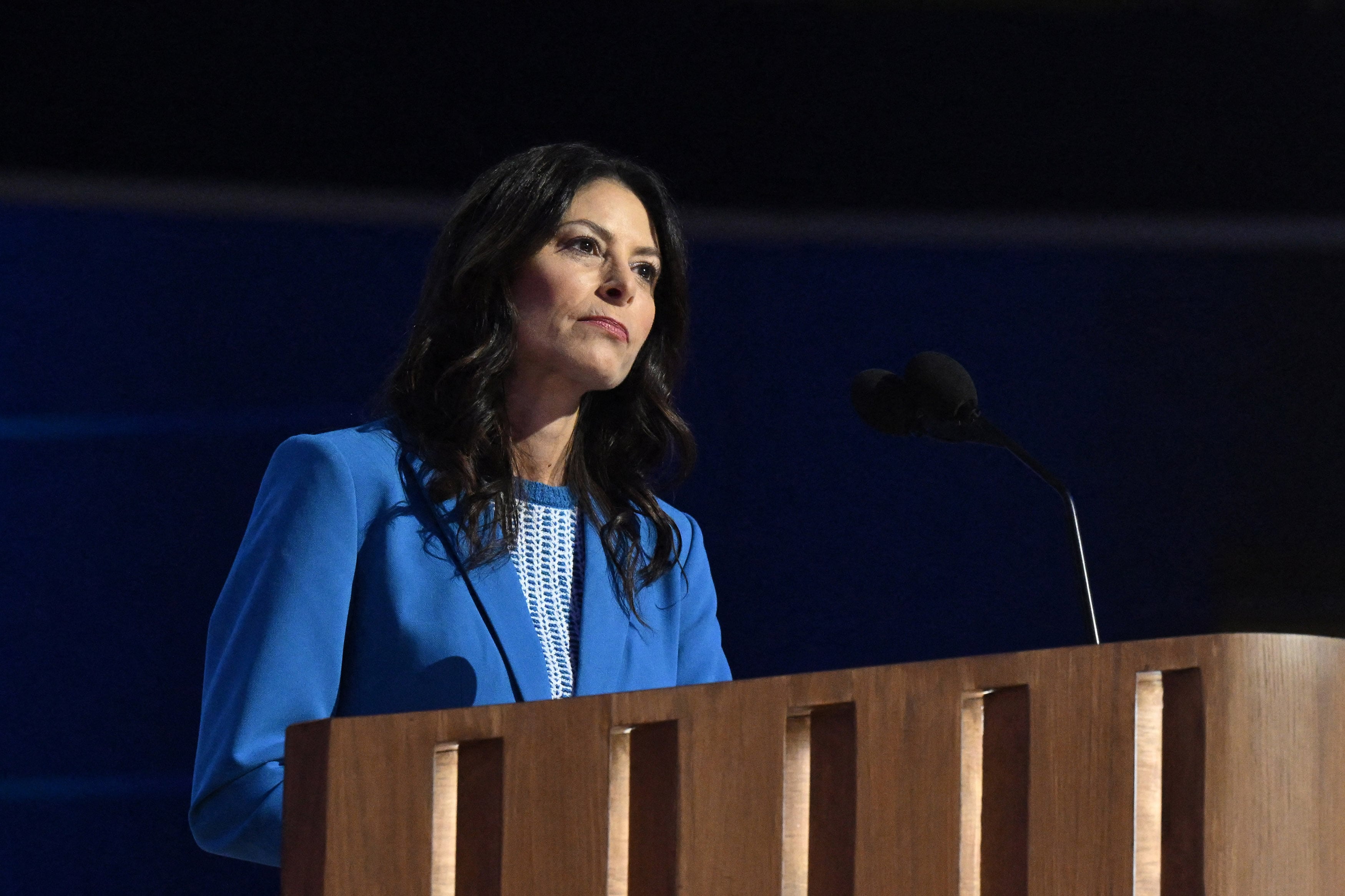 A white woman with long dark hair wearing a blue suit speaks from behind a wooden podium.