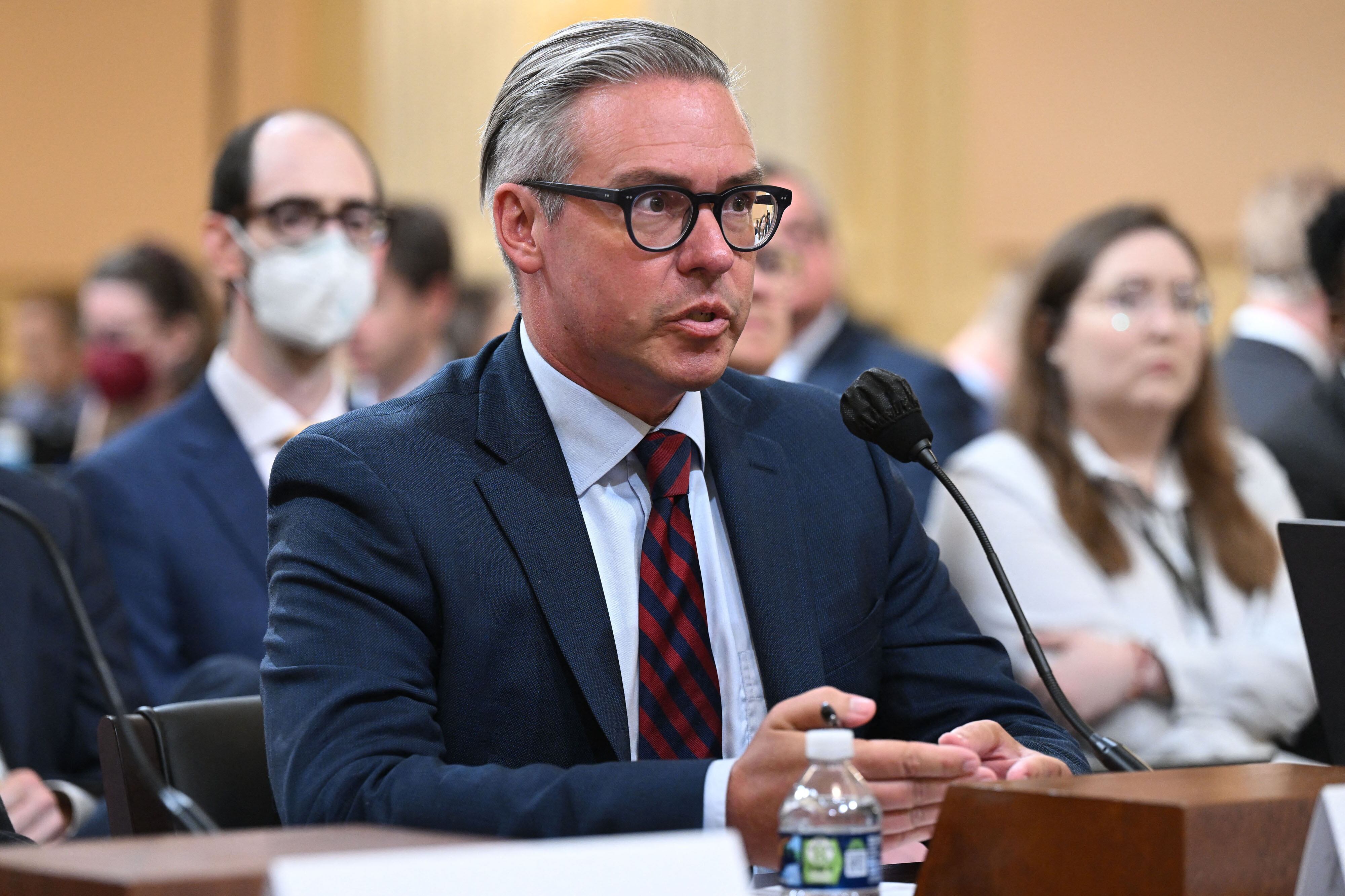 A man wearing glasses and a dark suit speaks from behind a microphone while sitting at a wooden desk with people sitting behind him in the background.