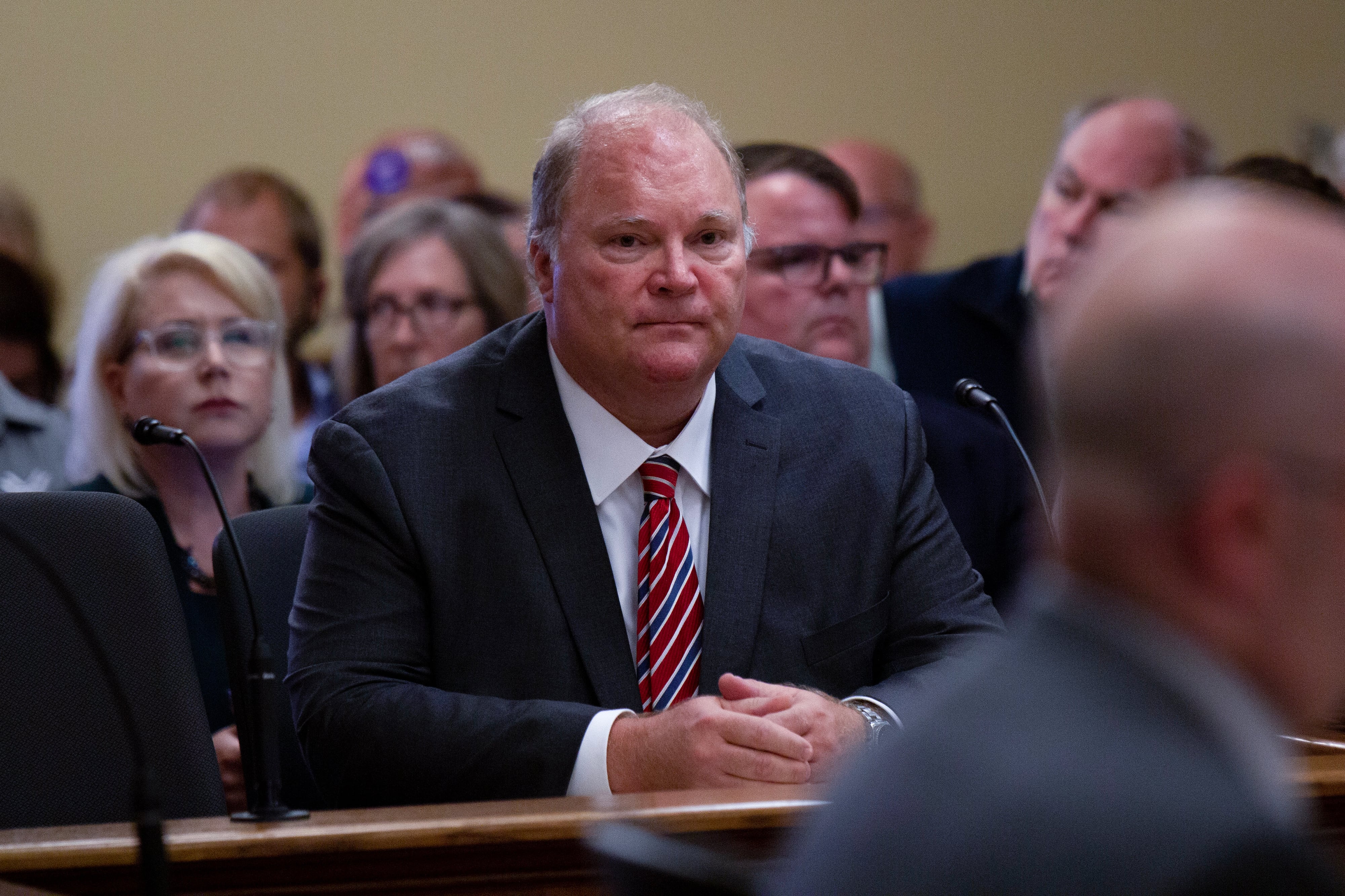 A photograph of a white man in a suit sitting at a wooden table with people sitting in the background.