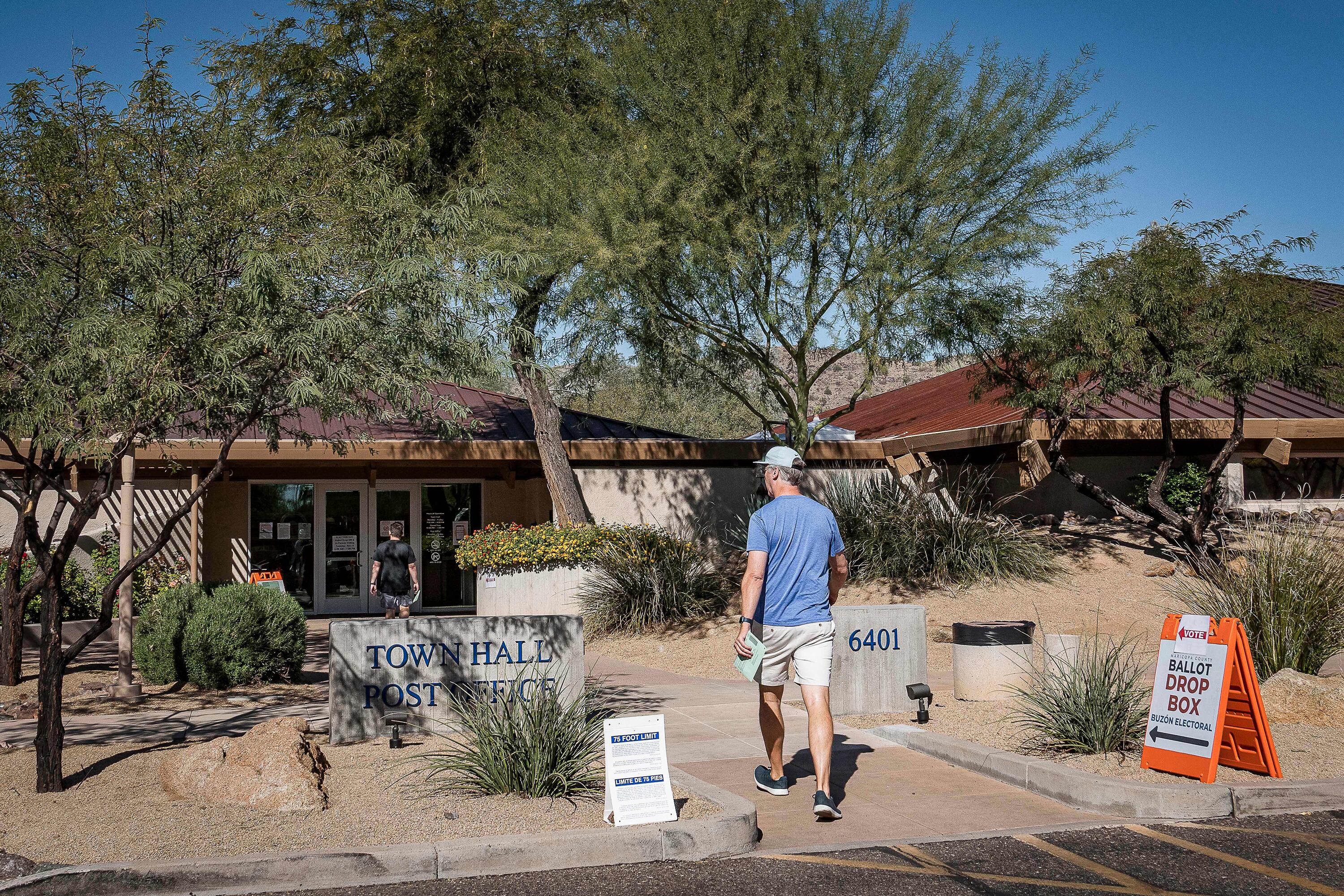 Two people walk toward the front entrance of a building with trees and signs in front of the building.