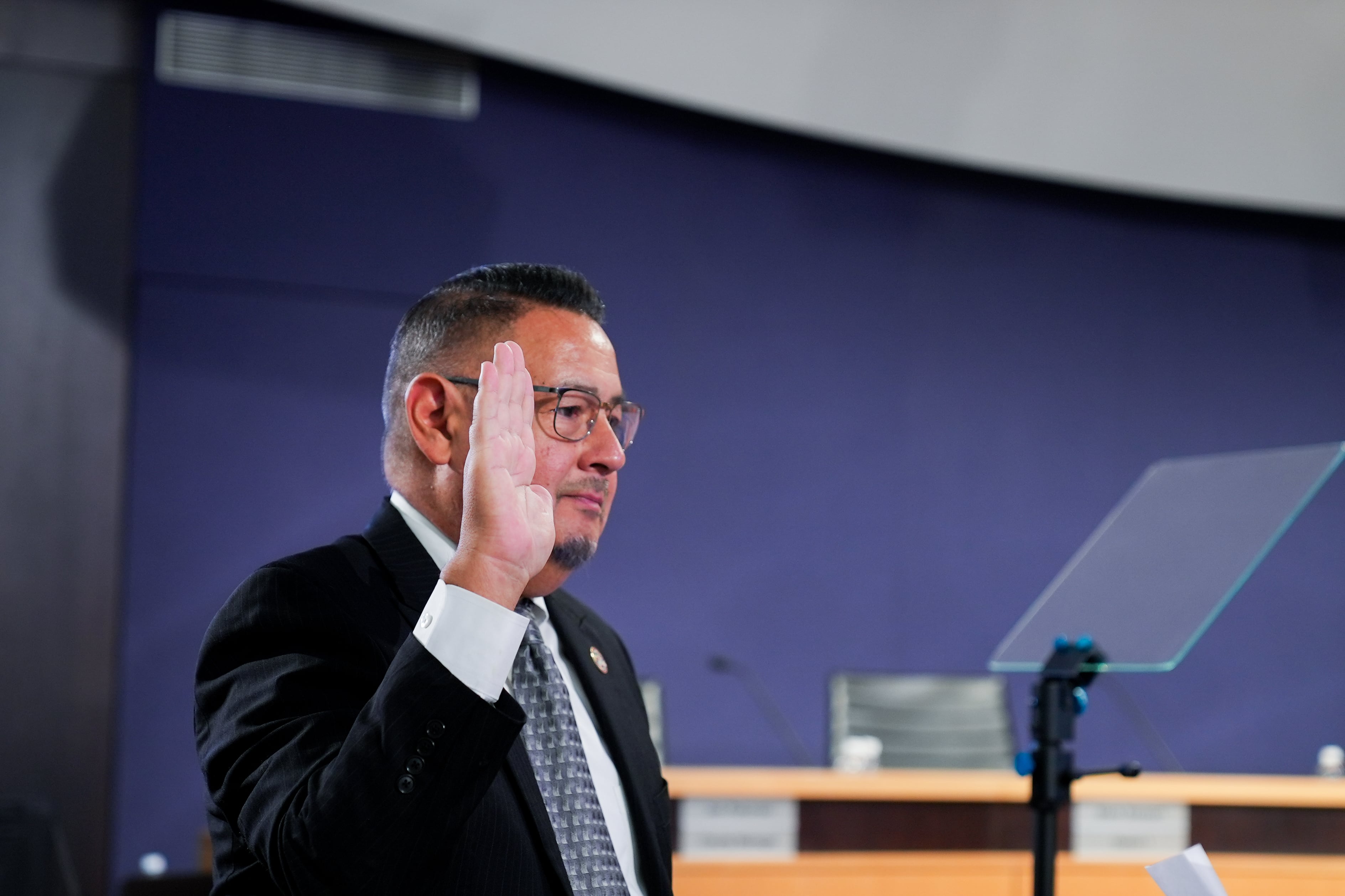A photograph of a man in a suit holding his right hand in the air while taking an oath.