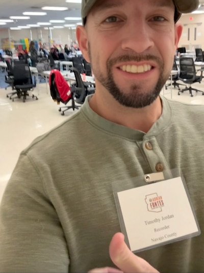 A screen grab of a man in a green shirt and wearing a baseball cap holding up his thumb and smiling.