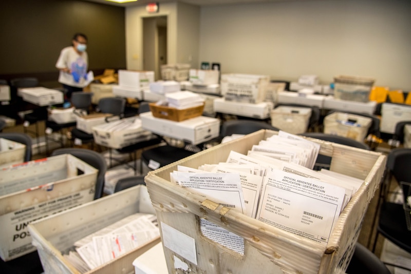 A room full of boxes full of ballots while a person wearing a mask stands in the background.