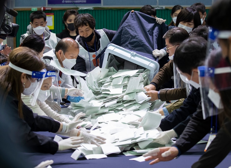 A large group of people in masks and all wearing gloves work to count ballots from a giant pile of ballots on a table.
