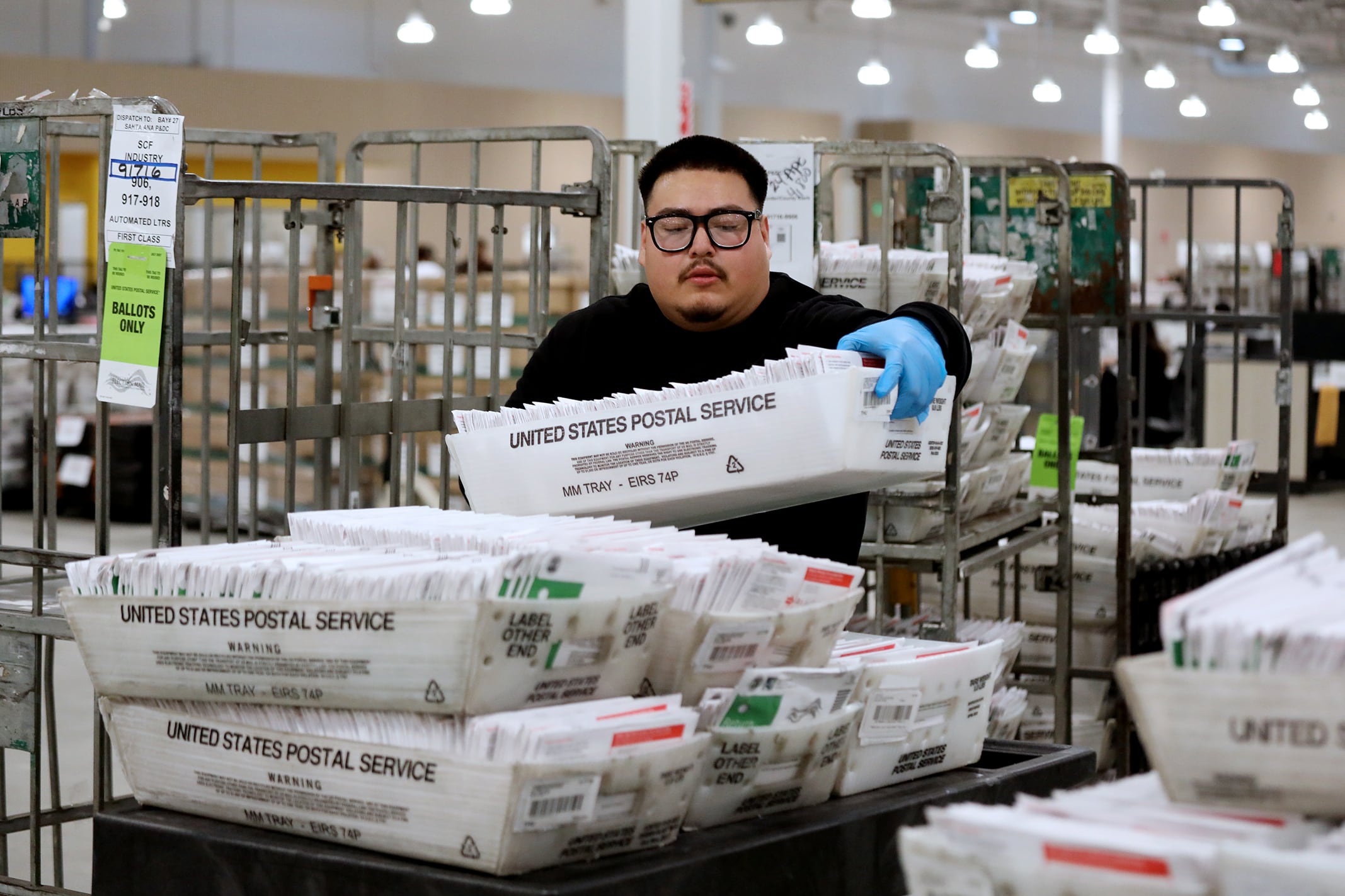 A man with dark hair, glasses and shirt holds a baskets of ballots at a Mail-in vote counting at Ballot Processing Center room.
