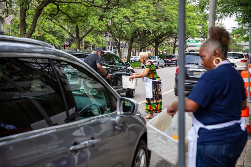 An adult woman wearing a white apron and holding a white box holds an orange envelope while standing next to a car with a window open while another woman wearing the same apron and holding the same box collects a ballot from a person in the stree.
