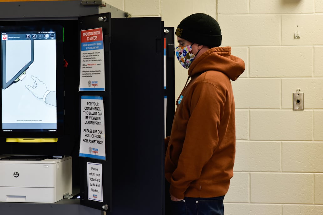 A man in a hoodie pictured in profiled in front of a voting machine, with a nearby screen showing instructions for voting