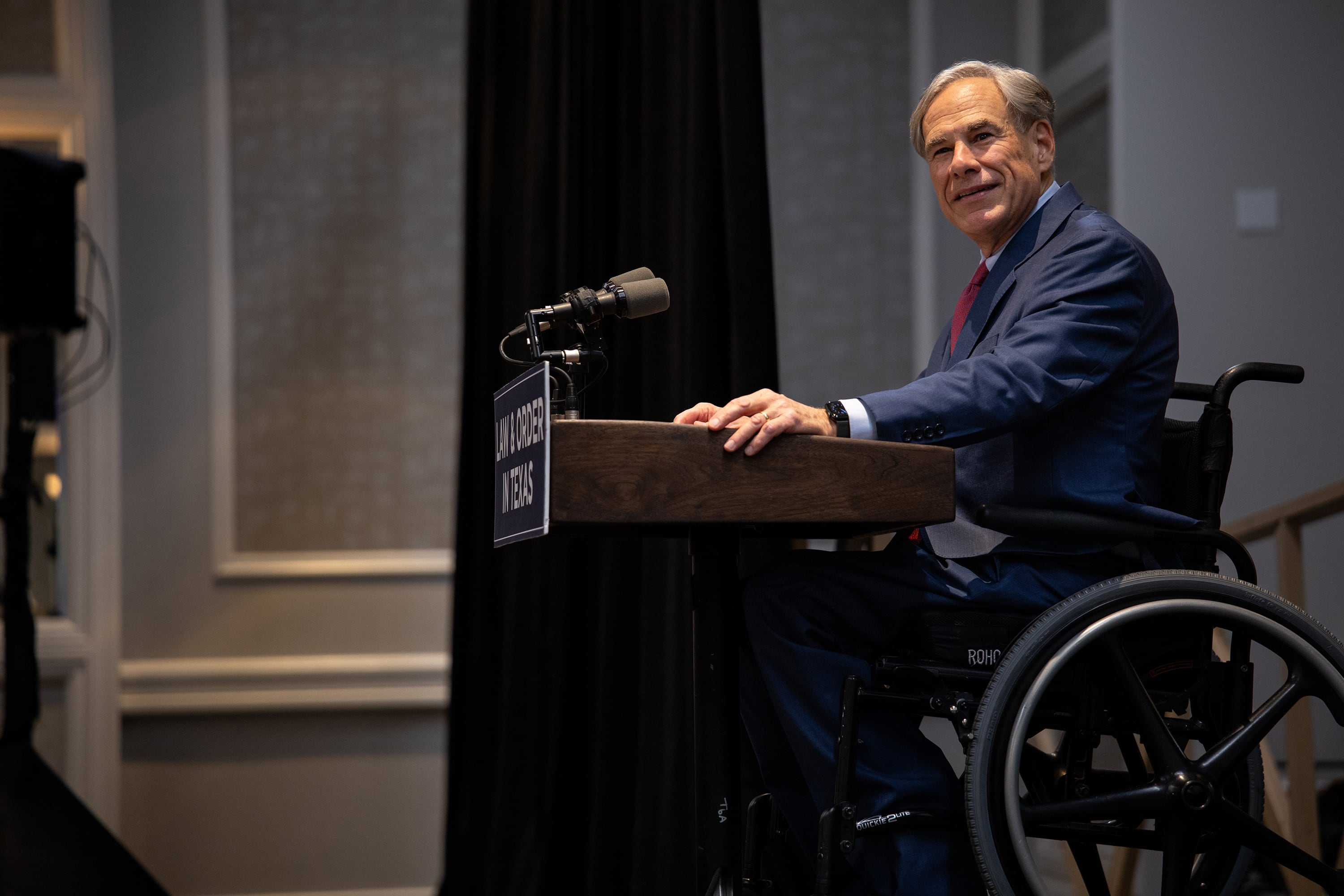 A photograph of an older white man in a suit and wheelchair sitting behind a podium.