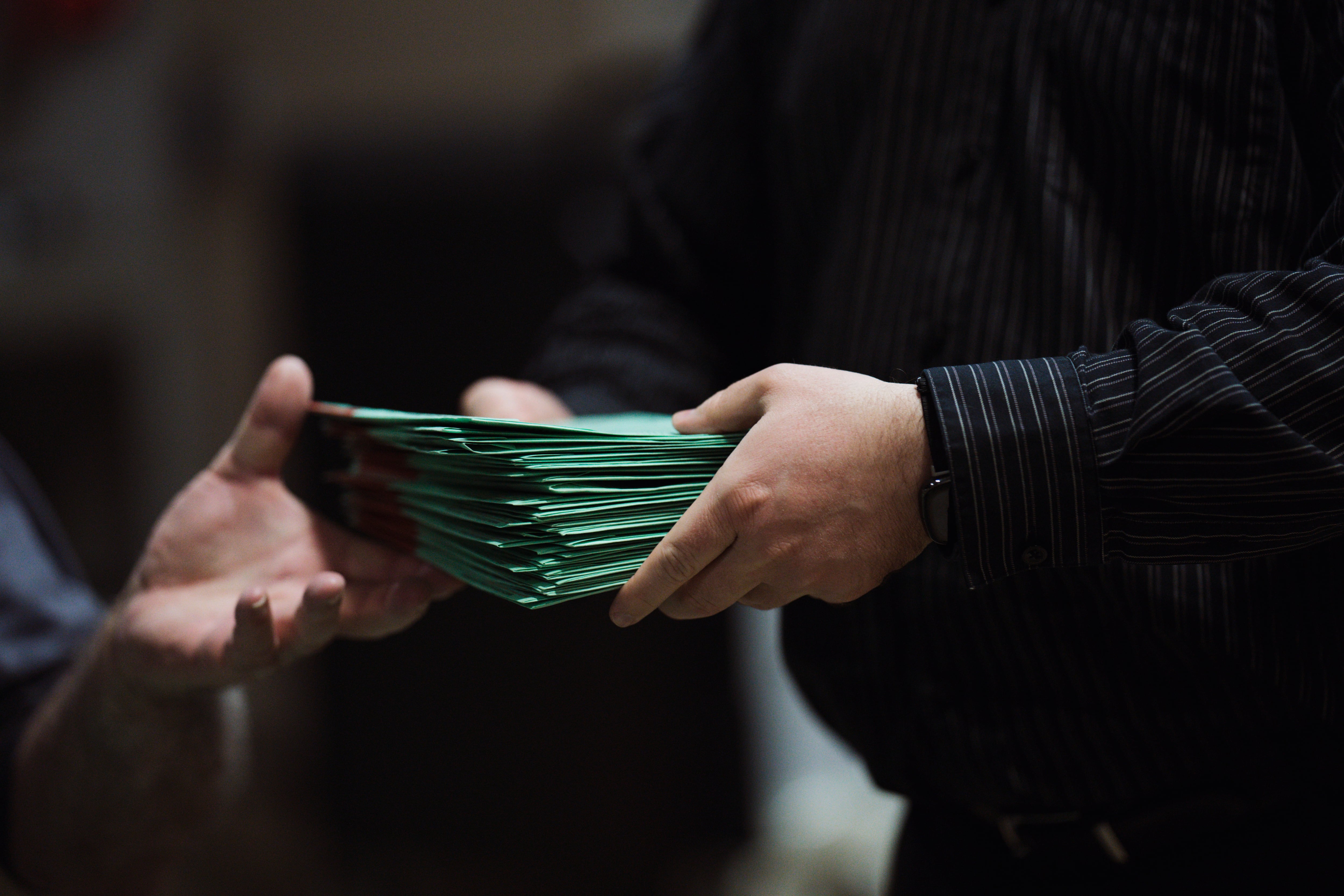 A closeup of a man's hands filled with a stack of green envelopes, passing them to another man's hand.