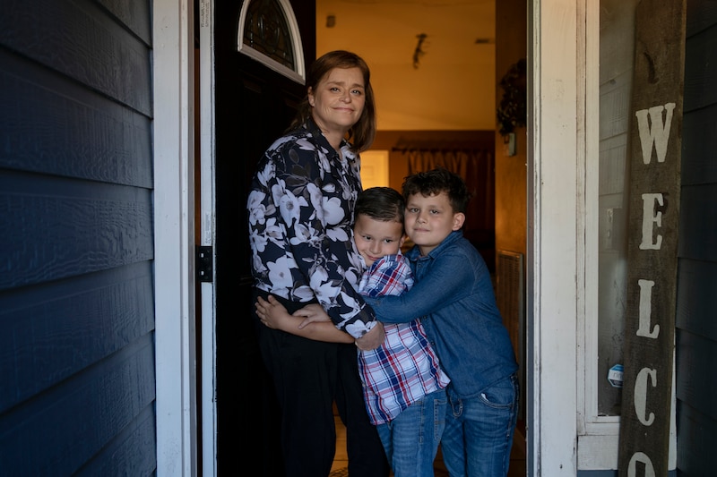 An adult and two young boys stand embracing each other in a doorway to their home.