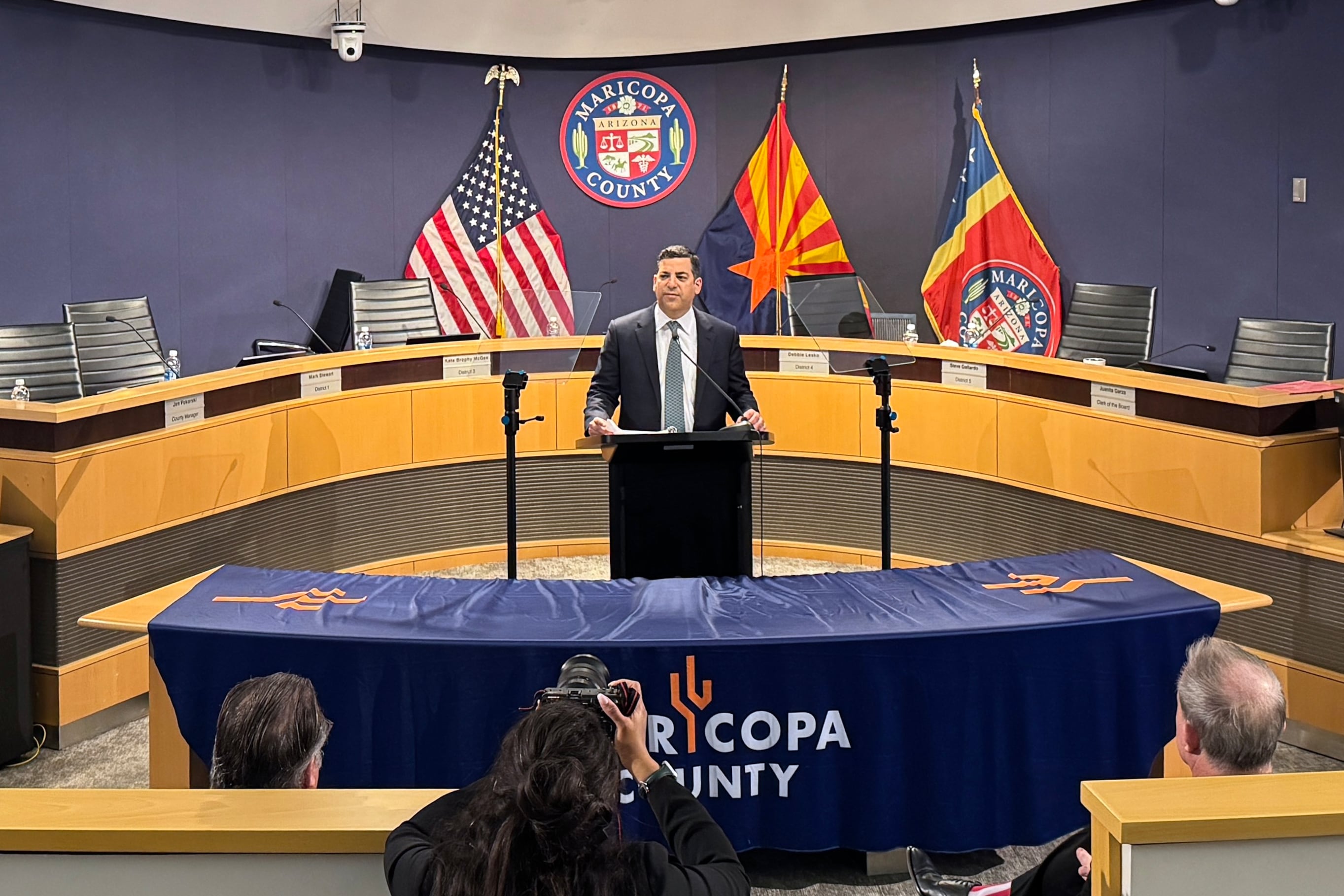 A man stands behind a podium with a long table and three flags in the background.