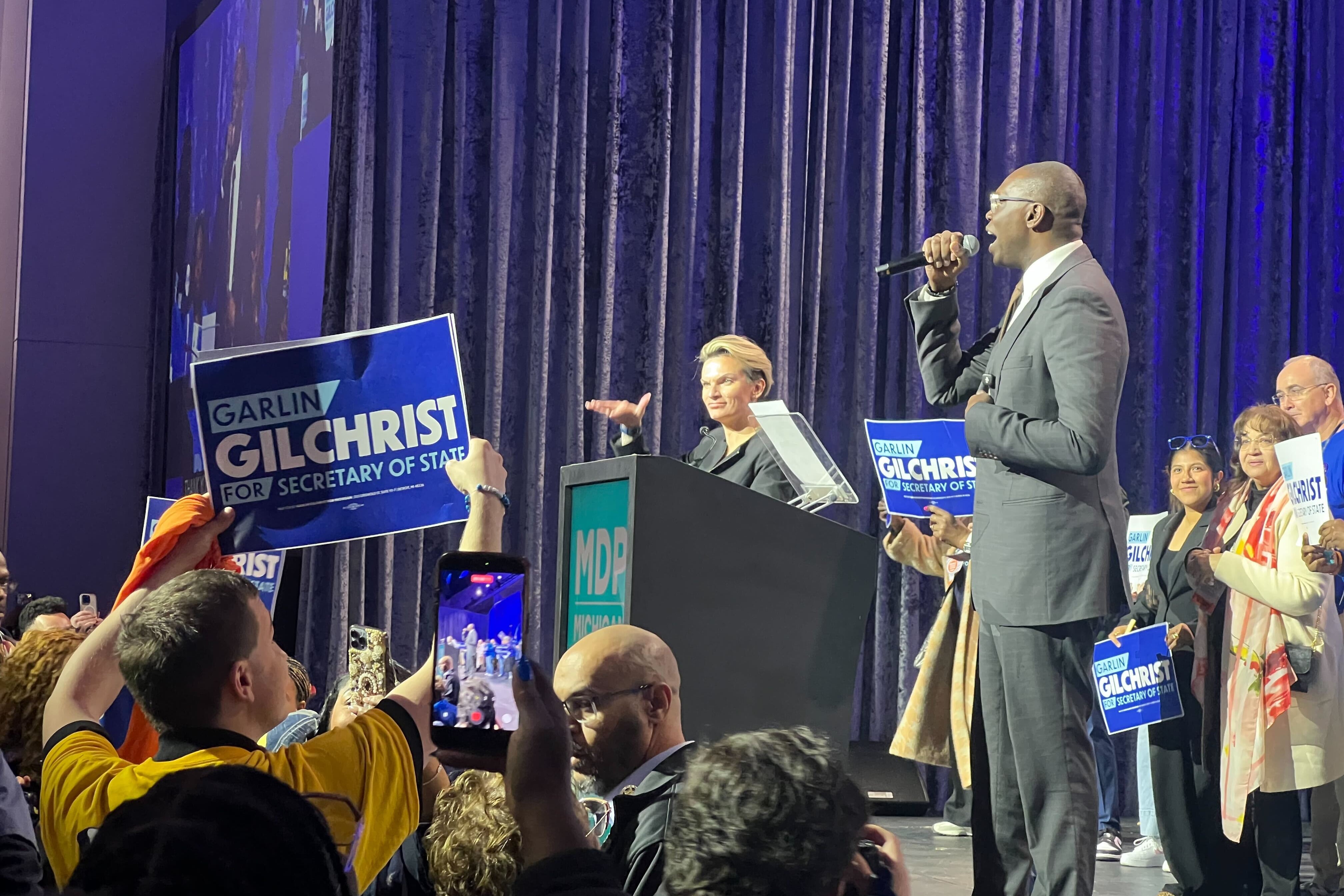 A tall man with a microphone on a dais speaks to a crowd holding up signs reading "Garlin Gilchrist"