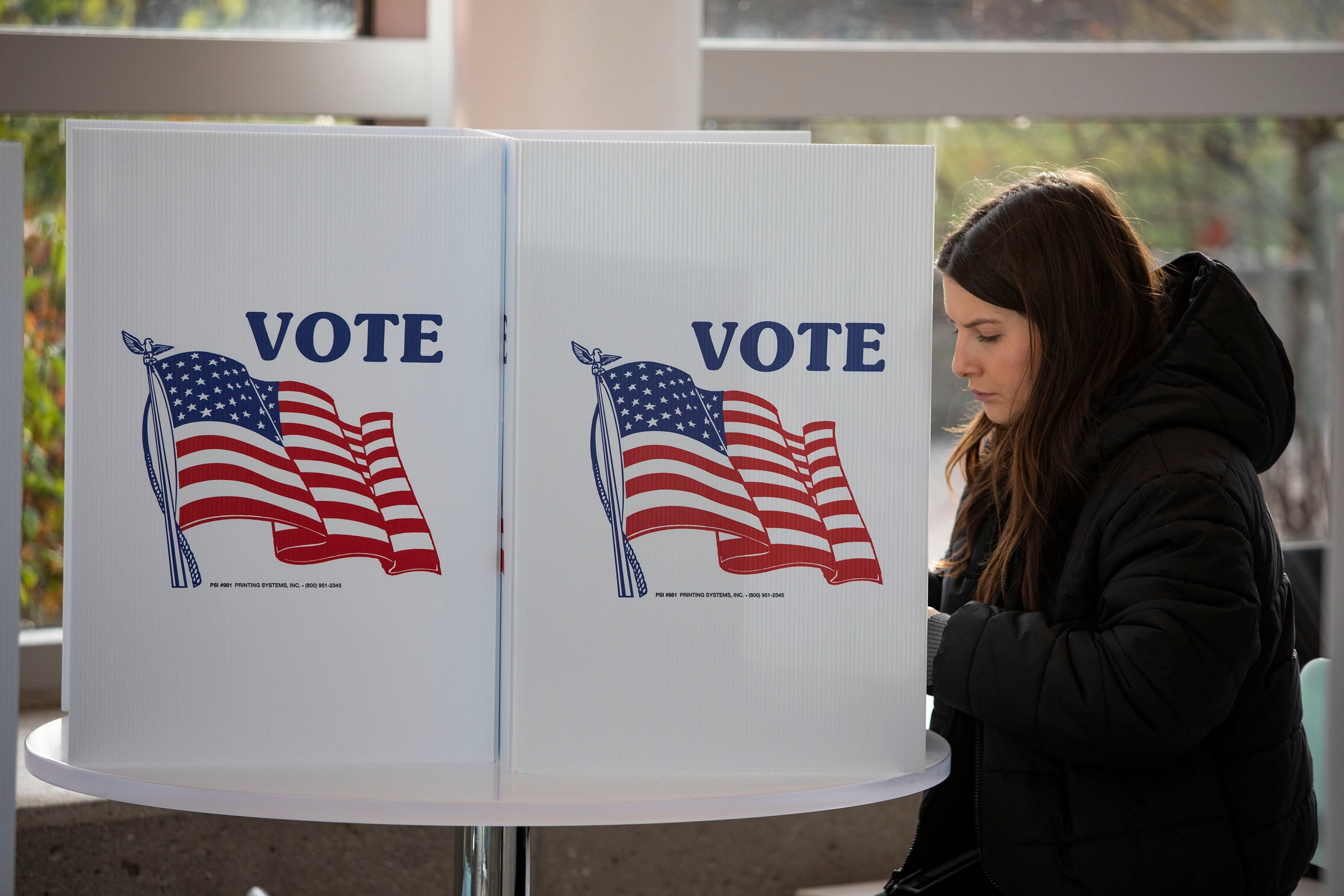 A young woman stands at a voting booth adorned with a large American flag and the word “Vote”