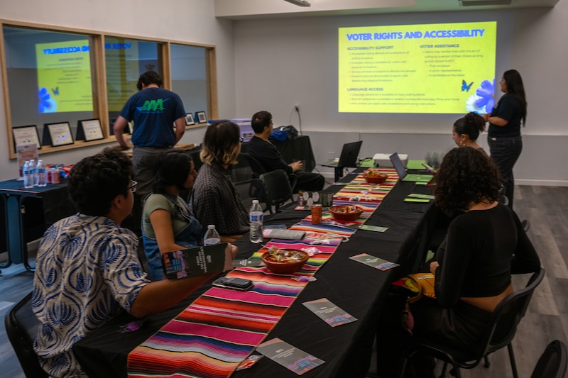A group of people sit around a long rectangular table while a person stands next to a screen at the end of the table and looking at a powerpoint.