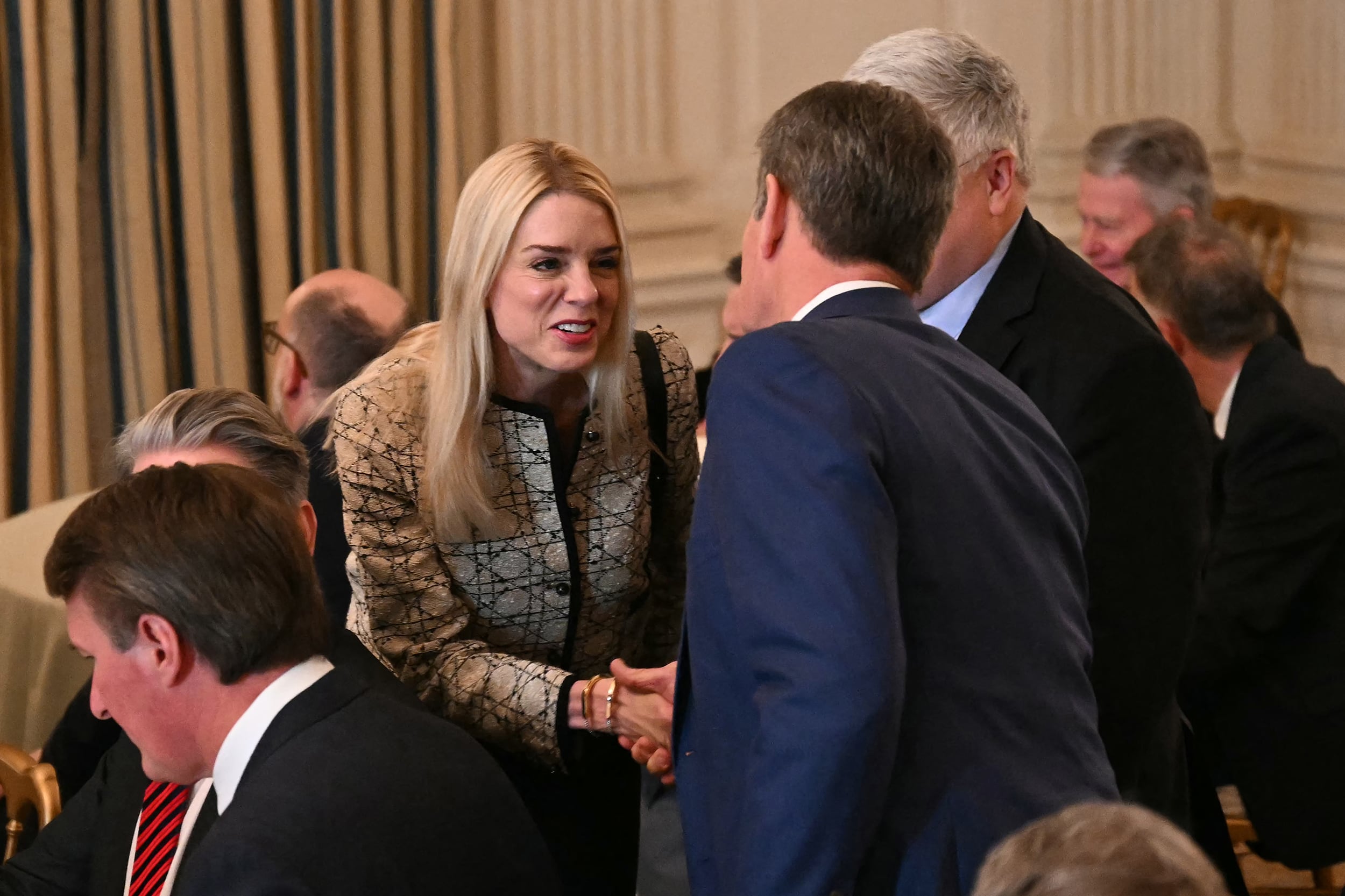 A white woman with blonde hair shakes the hand of a man in a blue suit facing away from the camera with other men in suits around them.