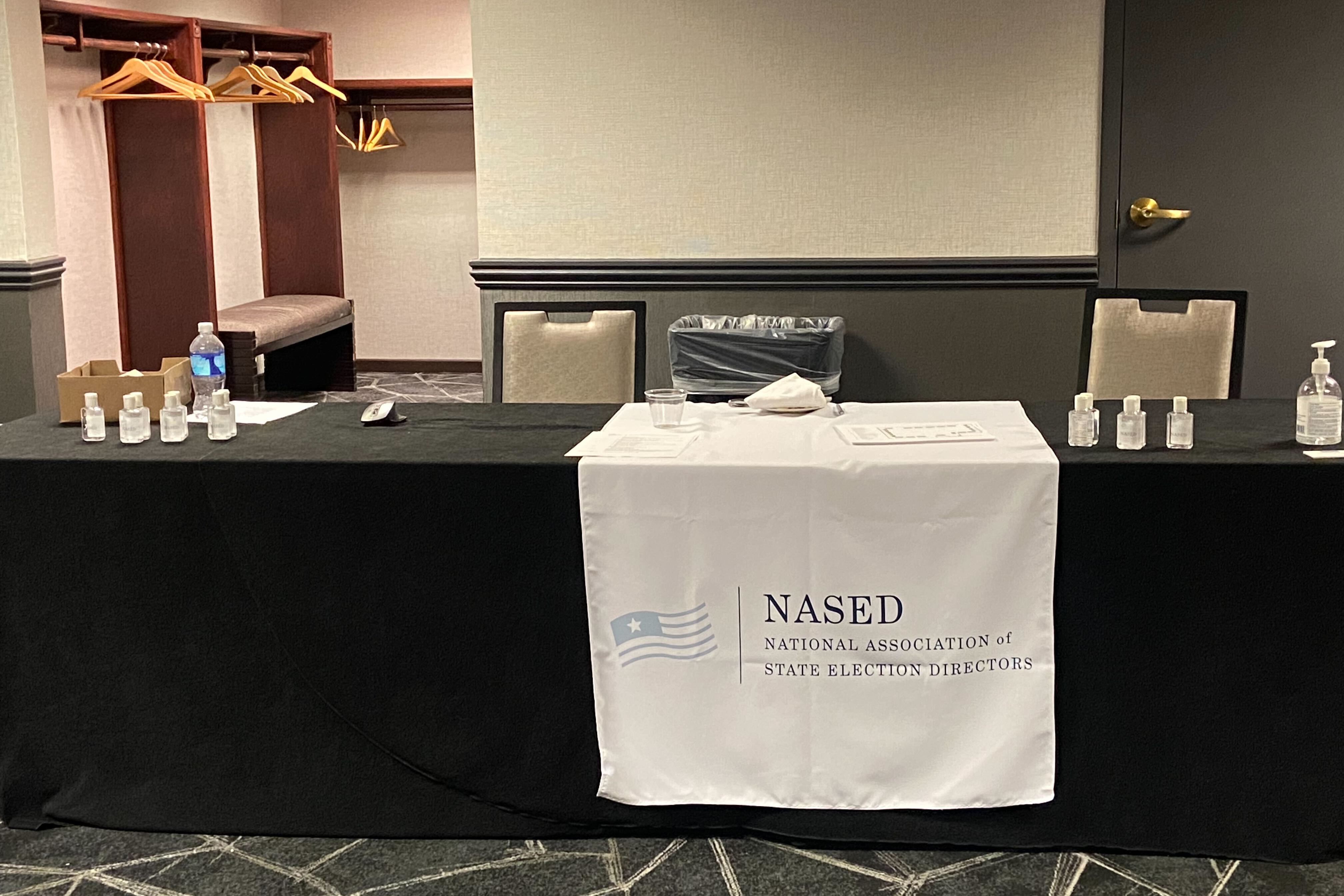 Water bottles and hand sanitizer sit on an empty table covered in a black tablecloth with a white banner reading “NASED - National Association of State Election Directors.”