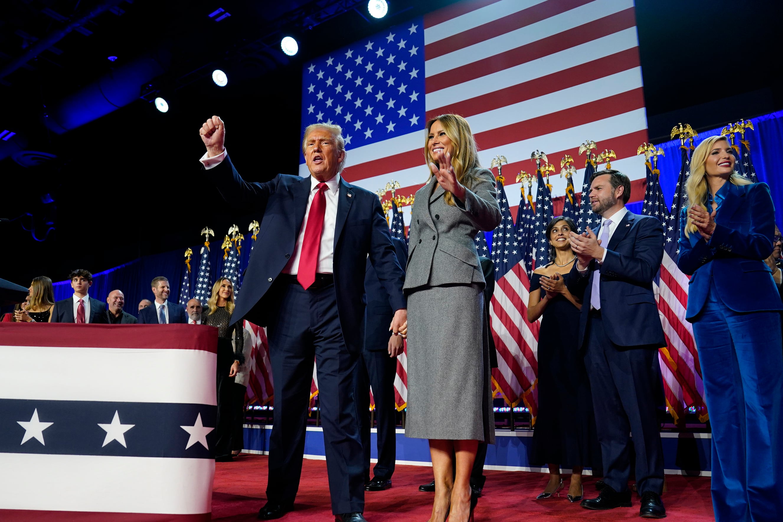 Two people stand on a stage with a wall of American flags in the background.
