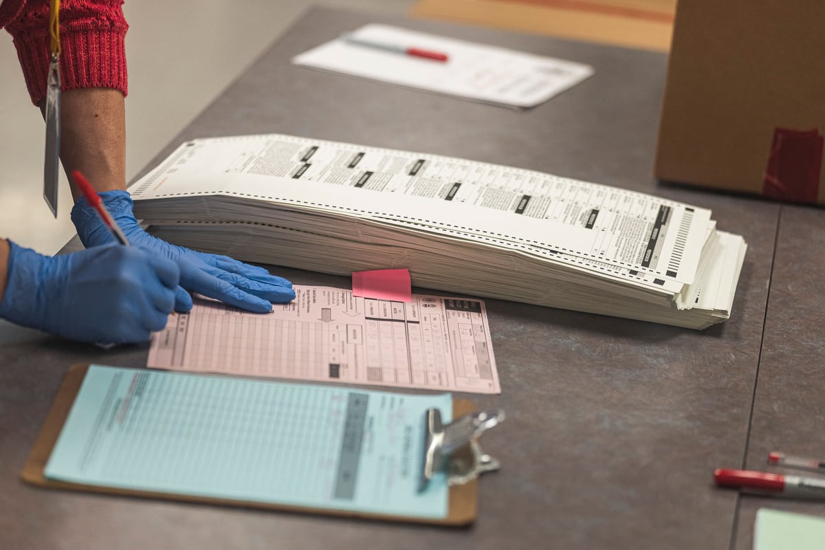 An arm sleeved in a red sweater and hands wearing blue disposable gloves holding a pencil and writing on a pink sheet of paper on a table between a pile of ballots and a clipboard with a blue piece of paper on it.