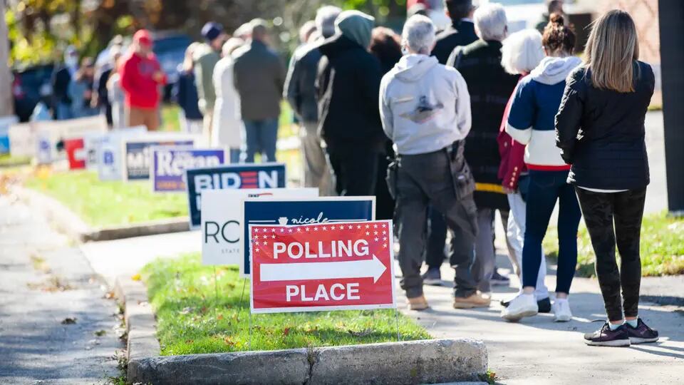 A long line of people on a sidewalk lined by campaign signs, including one red sign reading, “Polling place” with a large arrow pointing right