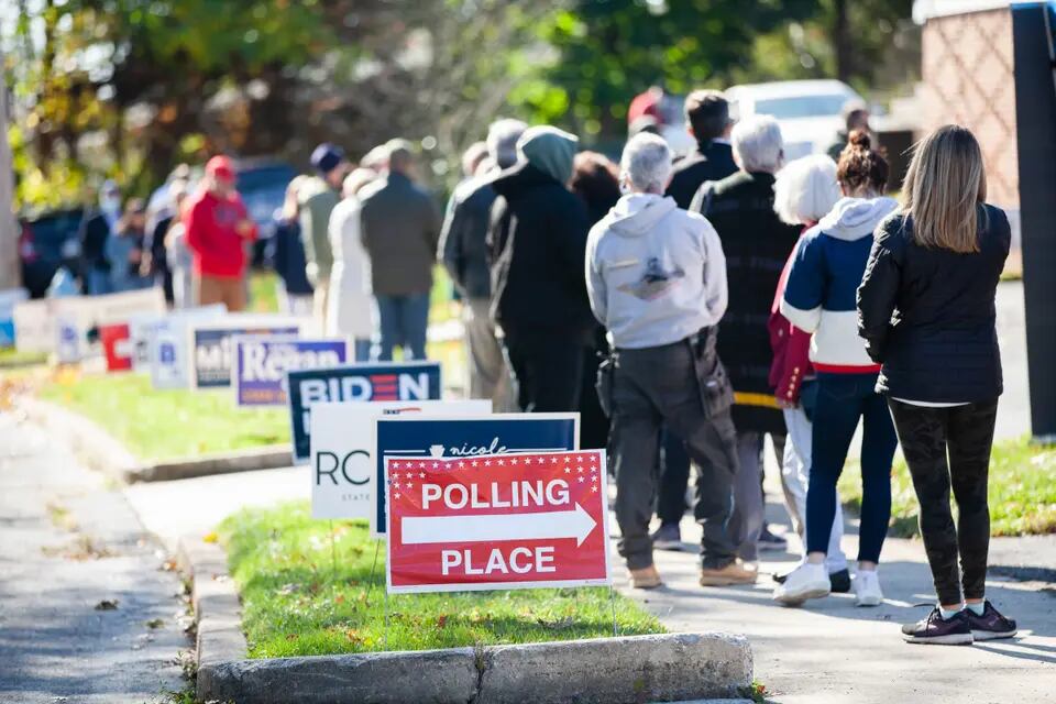 A long line of people on a sidewalk lined by campaign signs, including one red sign reading, “Polling place” with a large arrow pointing right