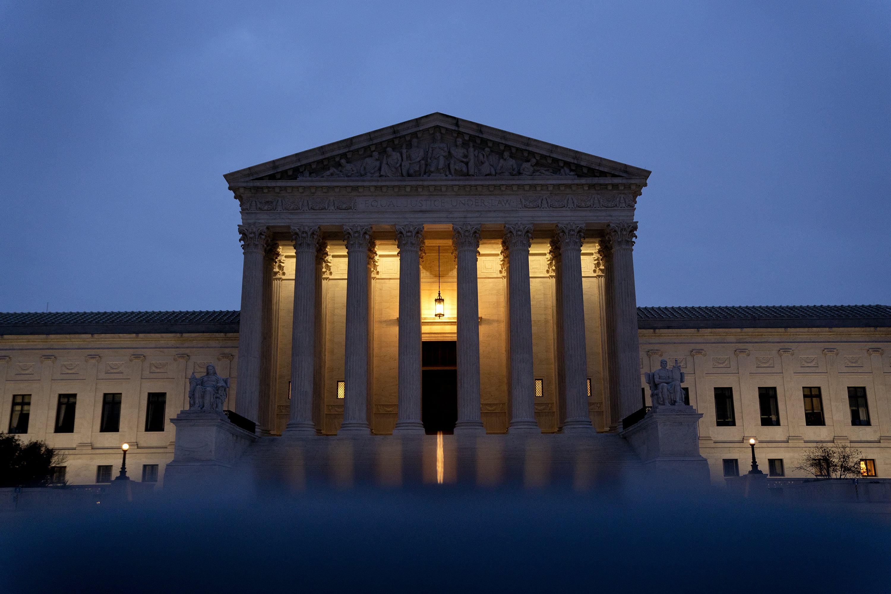 The U.S. Supreme Court building with large stone pillars at the front entrance with a dark blue sky in the background. The lights of the building are illuminating while the sun either sets or rises.