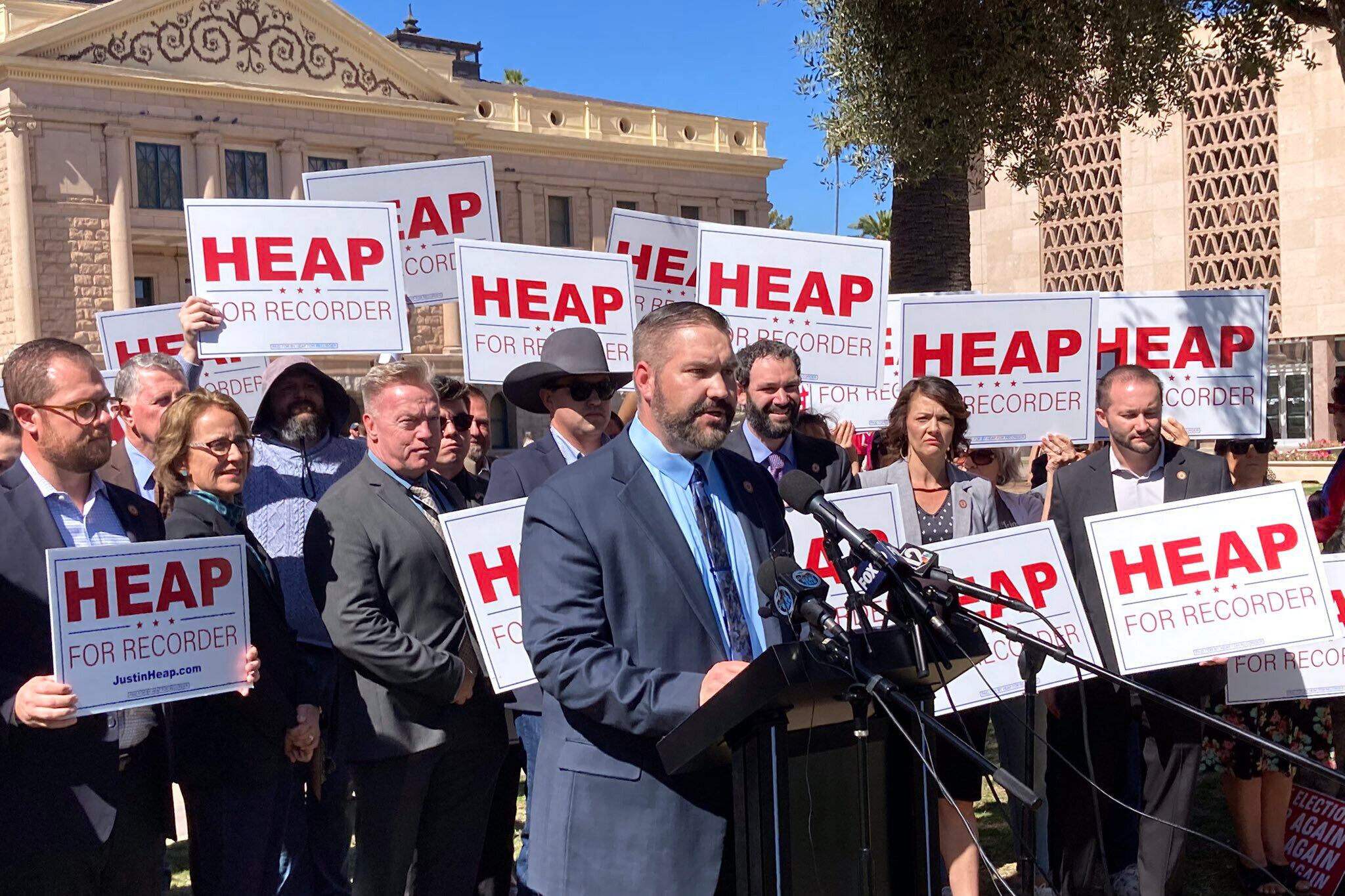 A group of people stand behind a man in a suit holding up red and white signs.