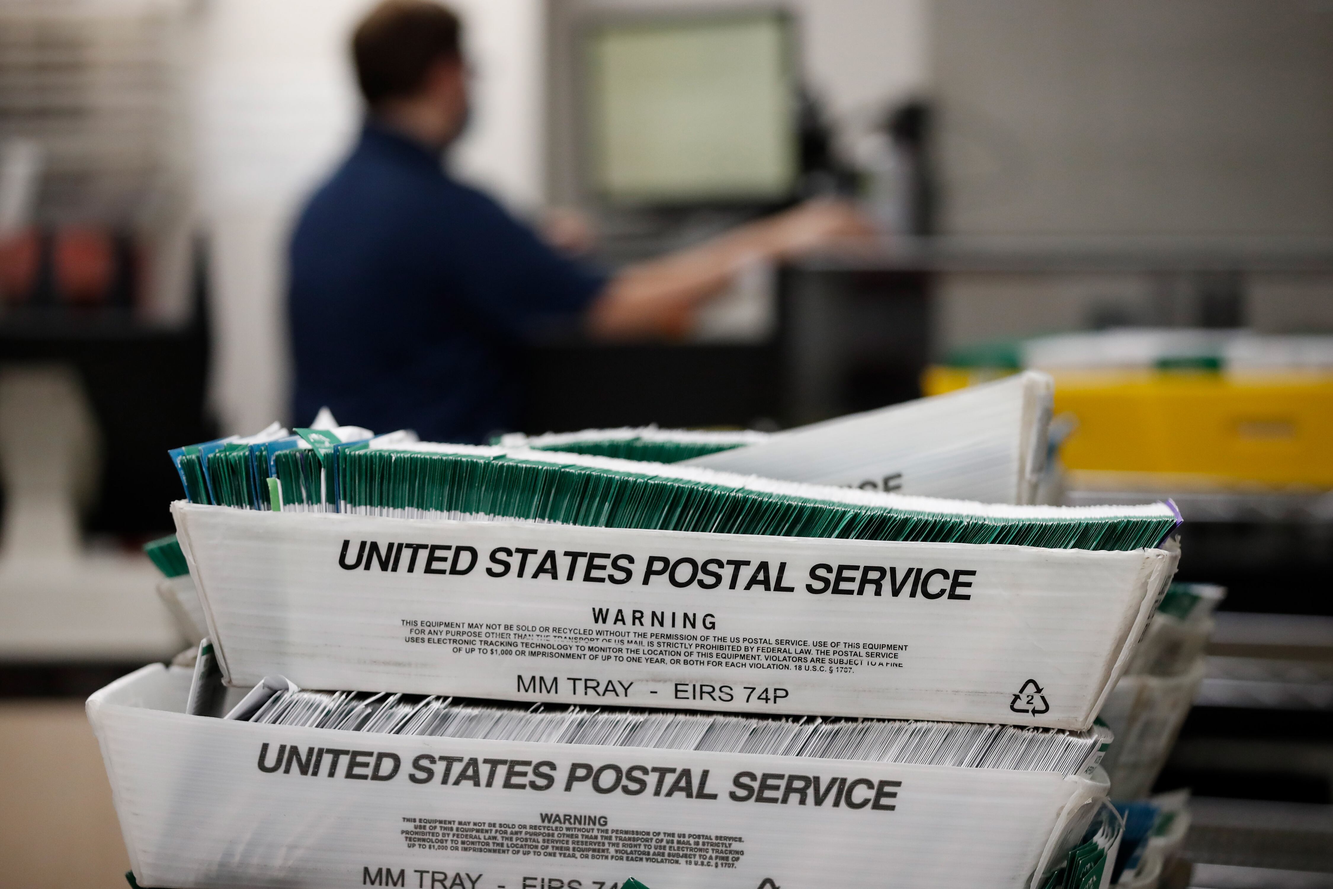 Ballots lined up in two white boxes stacked on top of each other and have black letters that read "United States Postal Service" with a person working in the background.