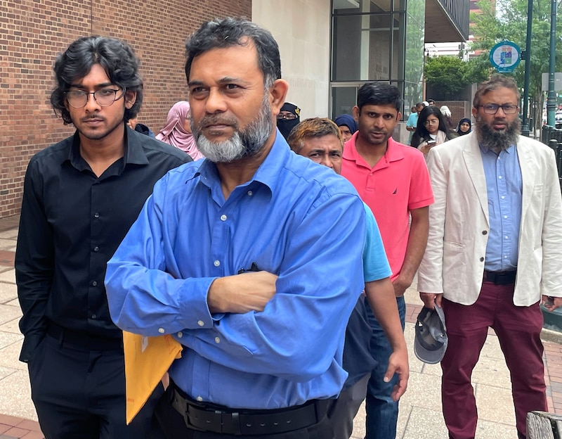 A man in a blue shirt walks with his arms crossed in front of other peopole.