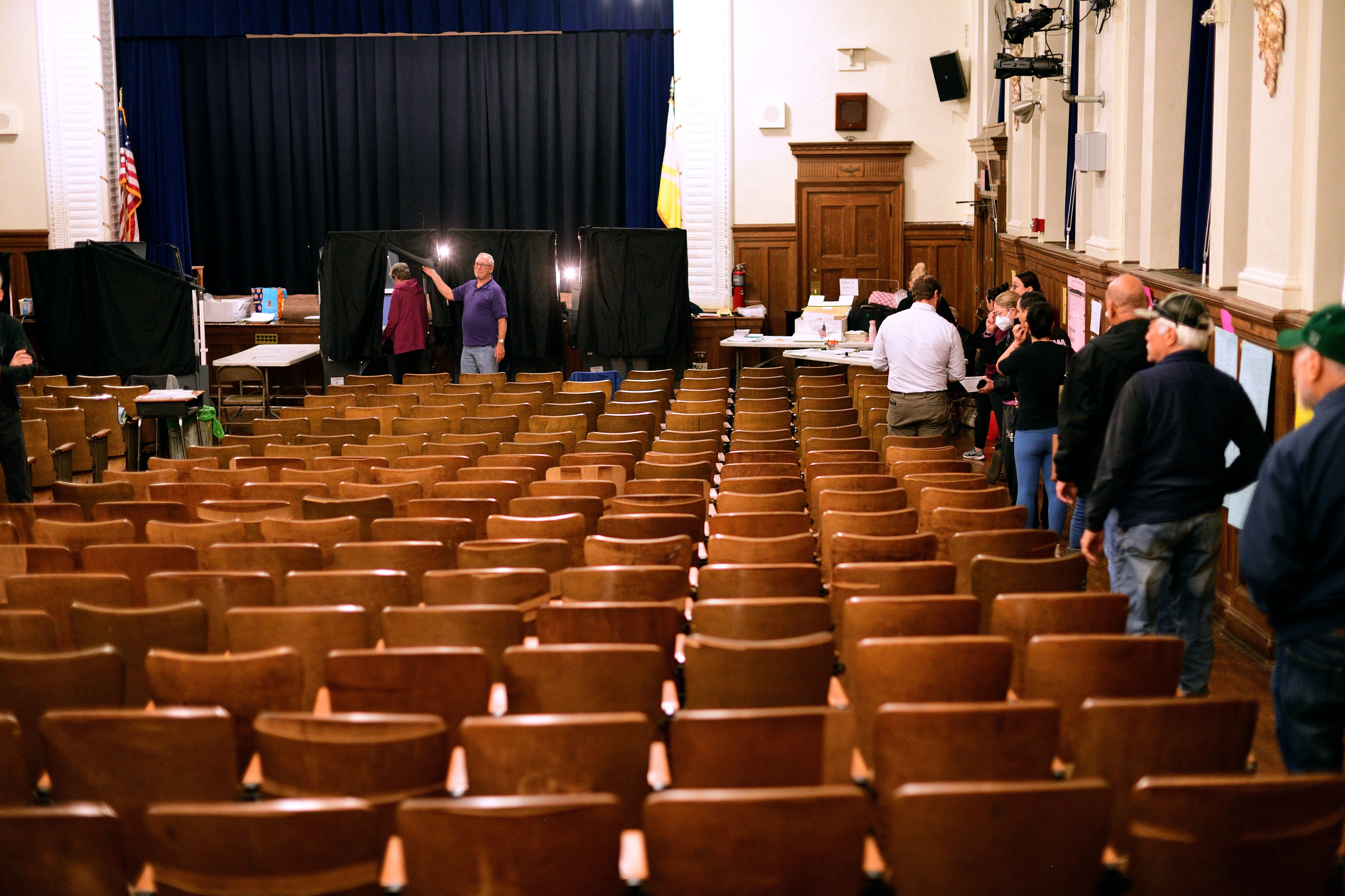 A line of people stand in a narrow aisle against a wall in a room filled with many rows of folding seats. A row of curtained voting machines stands at the front before a stage.
