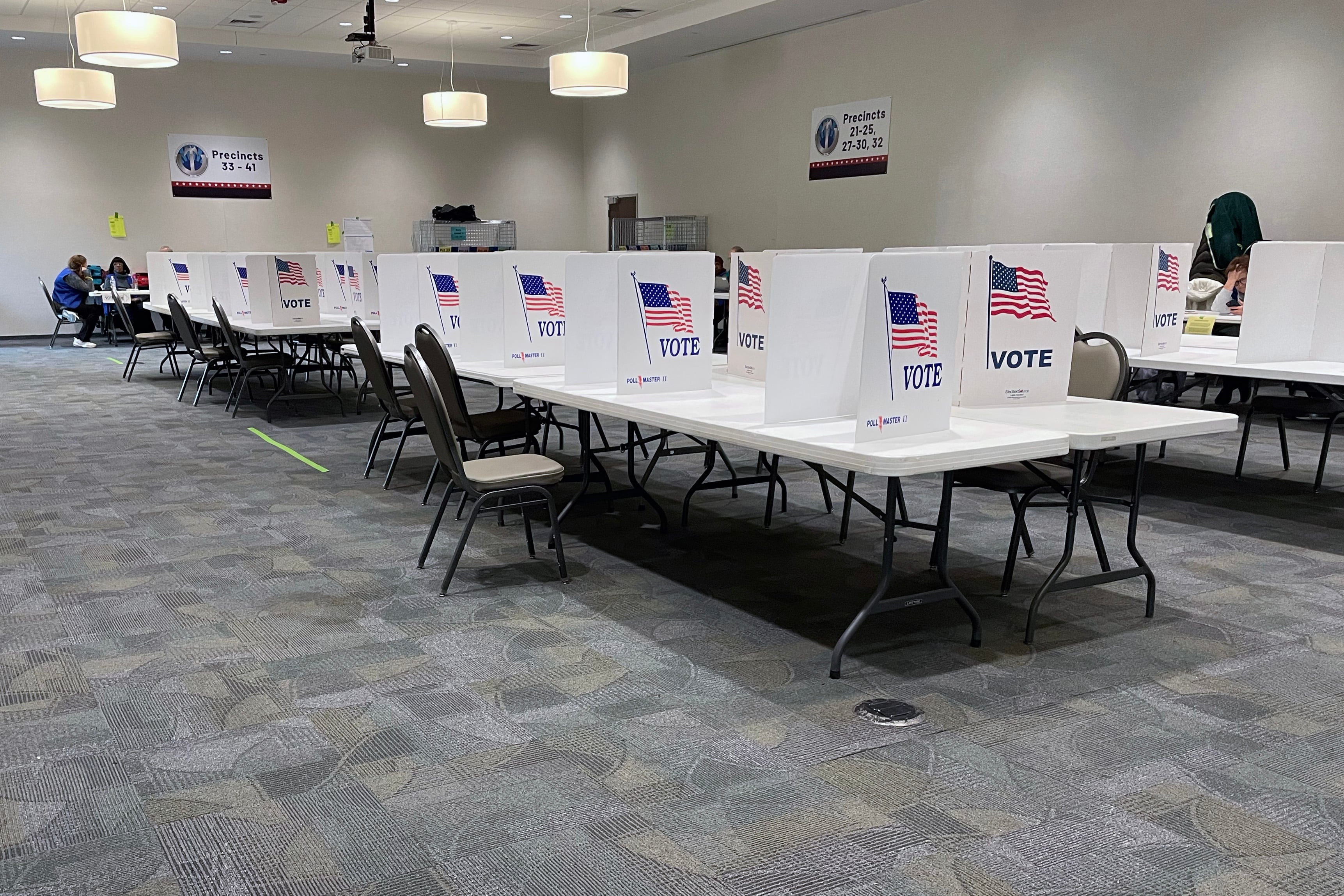 Cardboard dividers with the American flag and the word "Vote" line tables inside of a room with grey carpet.